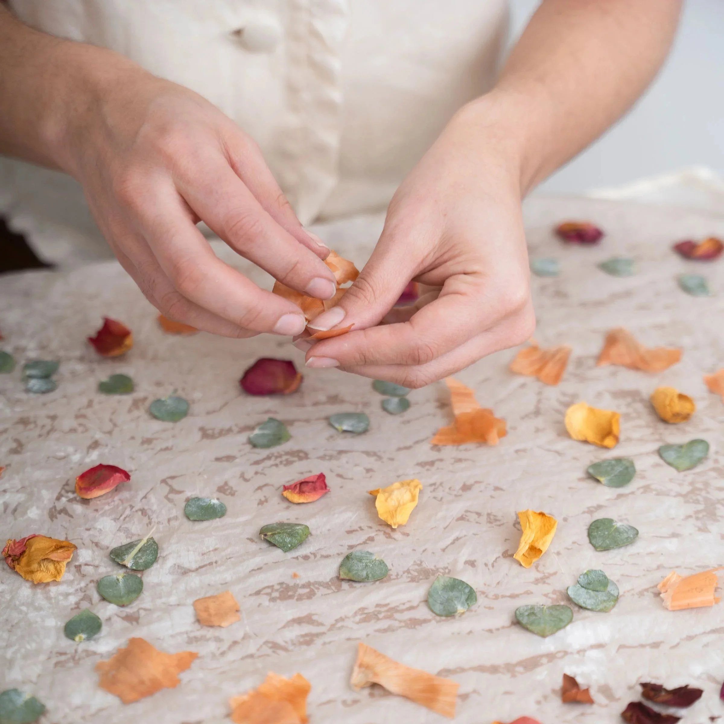 Hands peeling a dried orange peel over scattered dried orange and green leaves on a table.