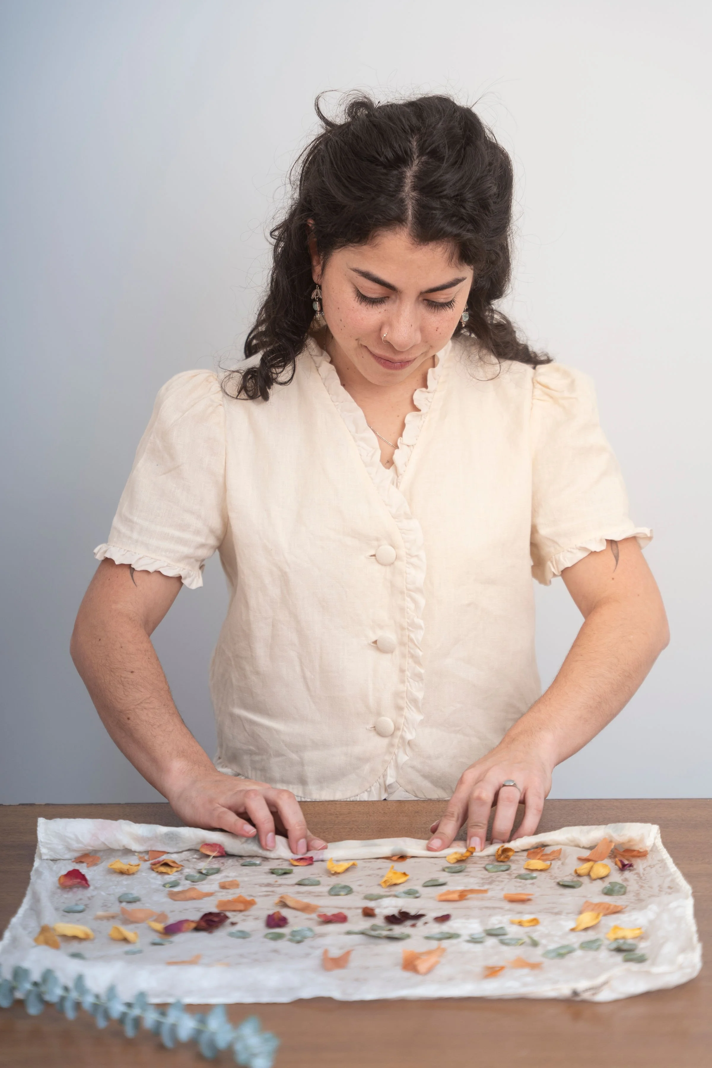 A woman with dark, curly hair and earrings, wearing a cream-colored blouse, is standing behind a table with parchment paper on it. She is arranging small, colorful, flower petal-shaped cutouts on the parchment paper.