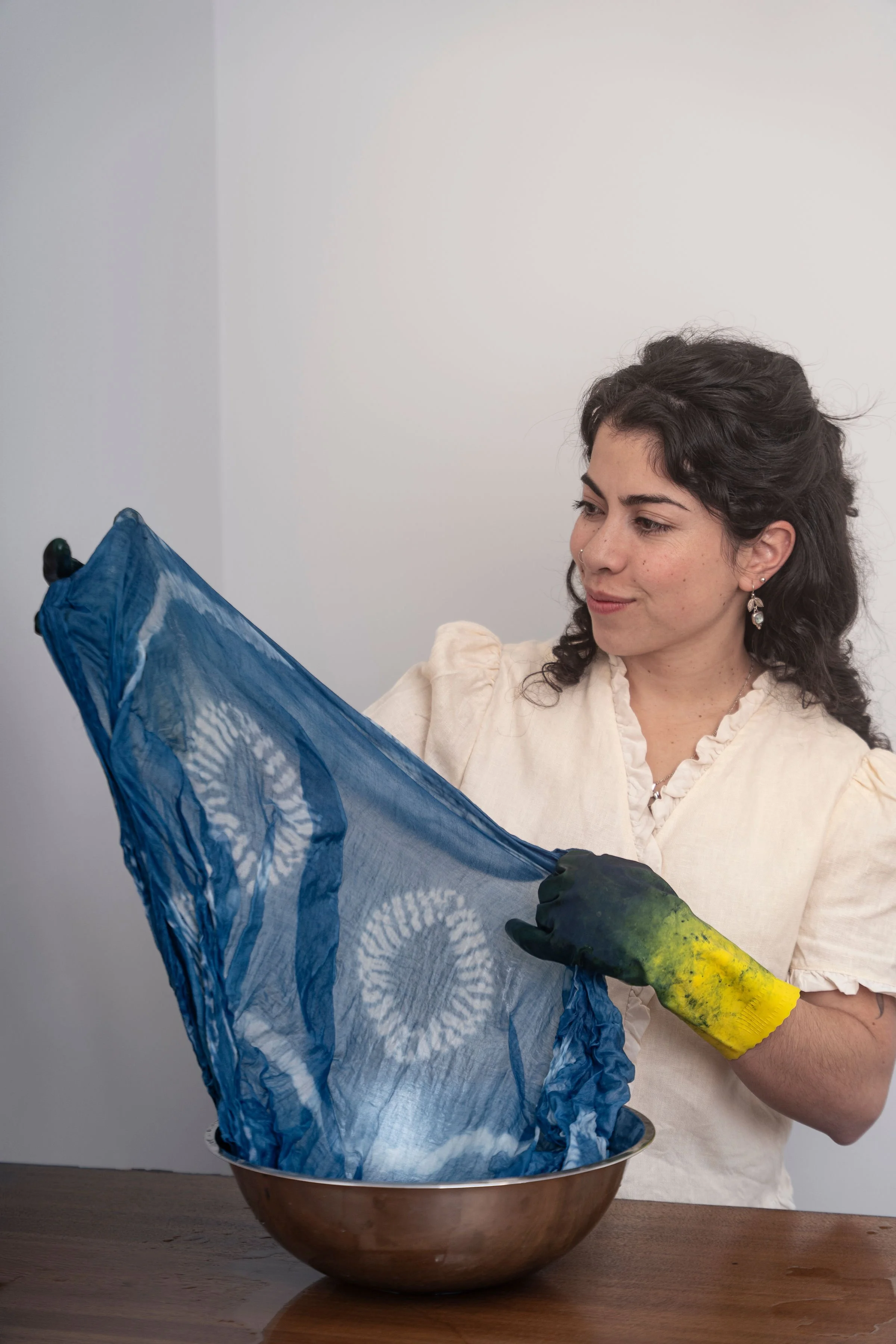 A woman with dark curly hair, earrings, and a pearl necklace is holding a blue fabric with white circular patterns and a green-gloved hand is holding a dye bottle above it, preparing to dye the fabric in a copper bowl.