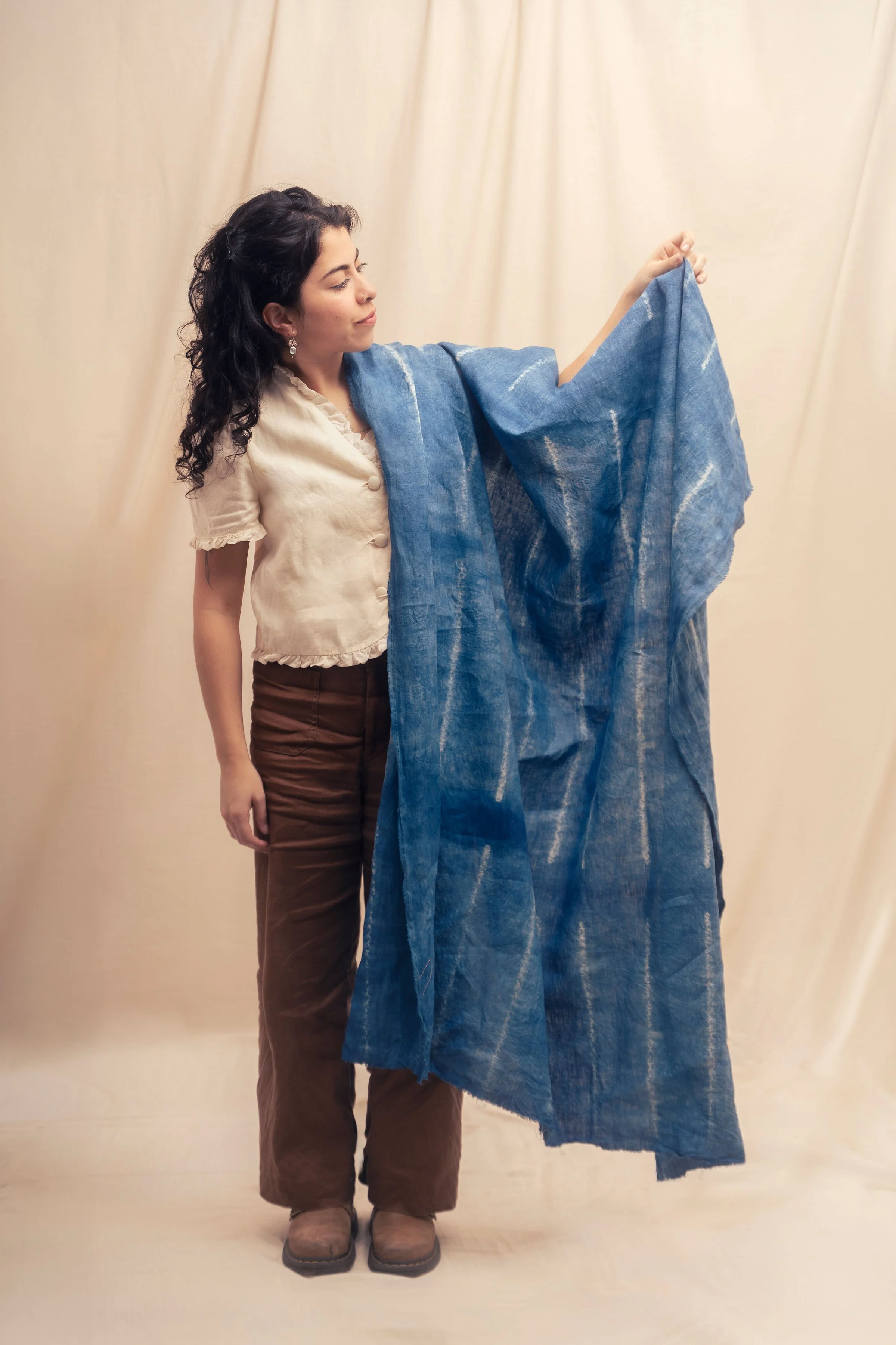A woman with dark curly hair, wearing a cream-colored shirt, brown pants, and tan boots, stands next to a large piece of blue fabric, holding it up with her right hand, in front of a beige curtain.