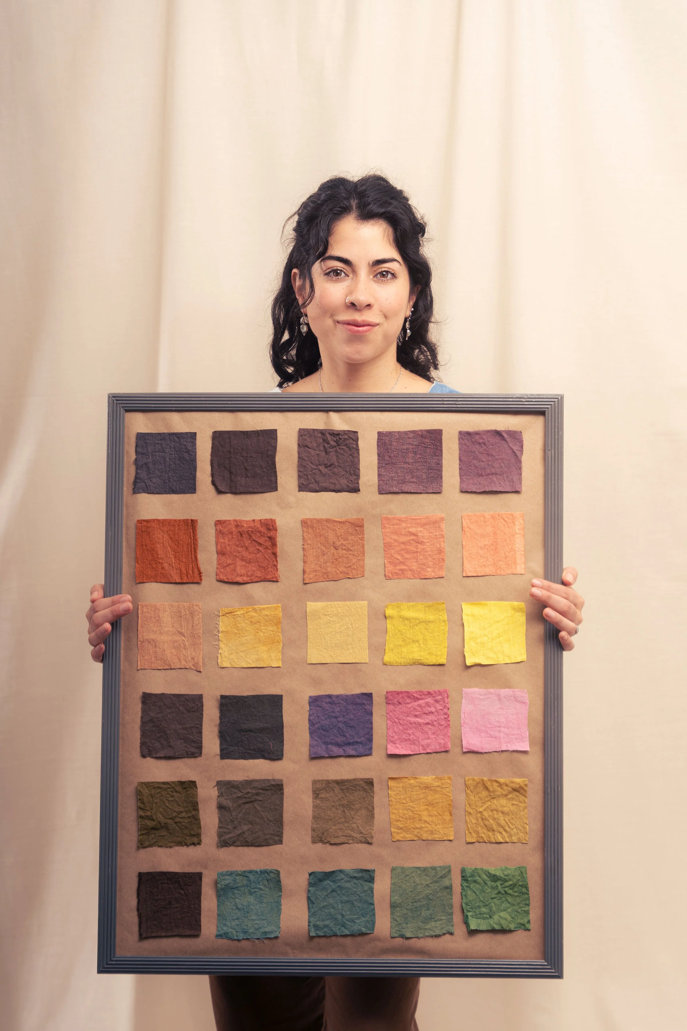 A woman with dark hair and earrings holds a framed display of colorful fabric swatches arranged in rows against a beige background.