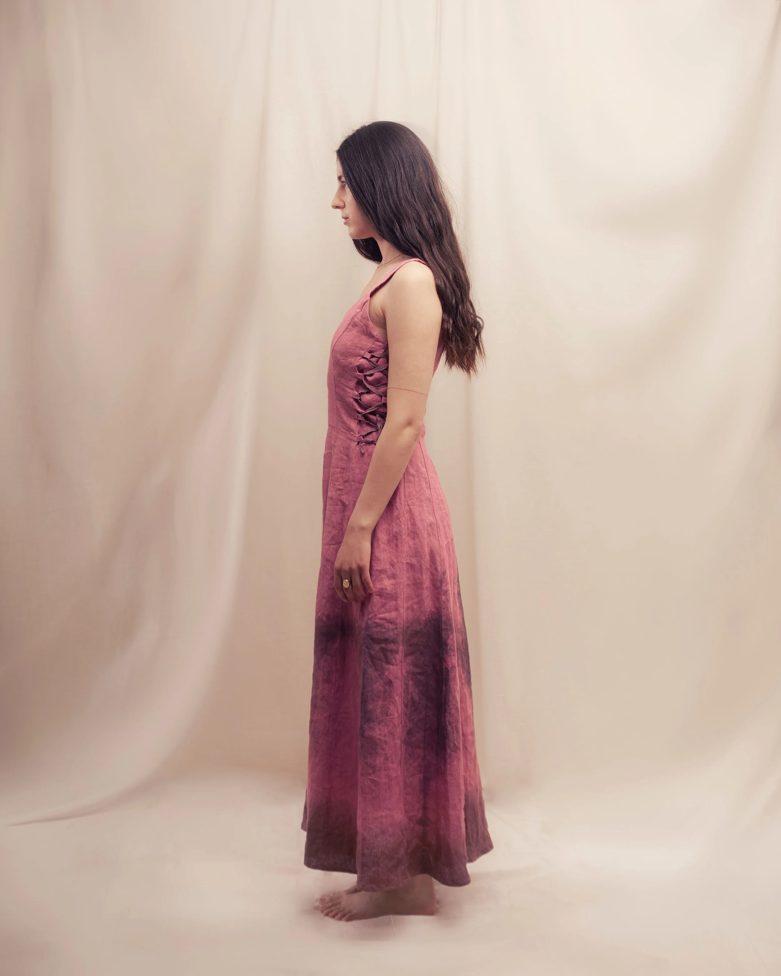 A woman with long dark hair wearing a sleeveless pink dress standing sideways against a neutral background.