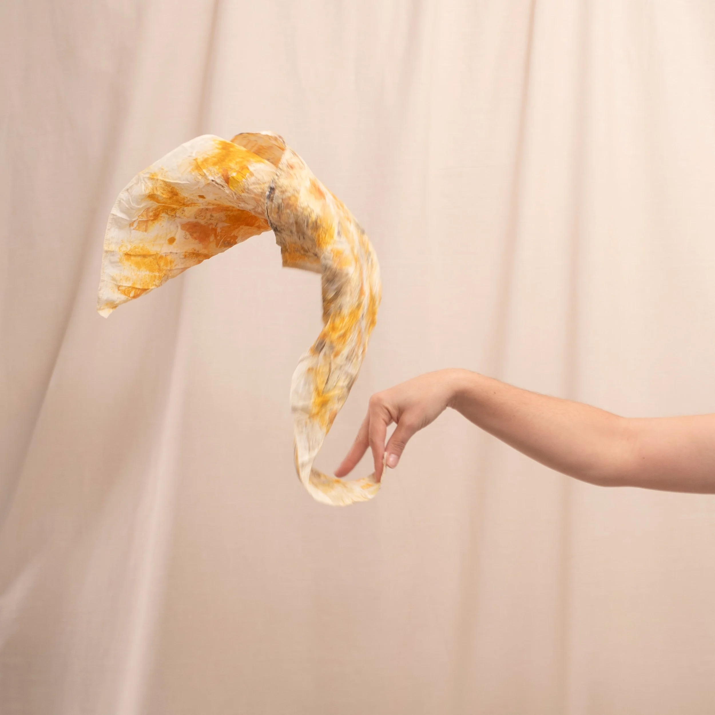 A person holding a crumpled piece of paper with orange, yellow, and brown paint marks against a beige curtain background.