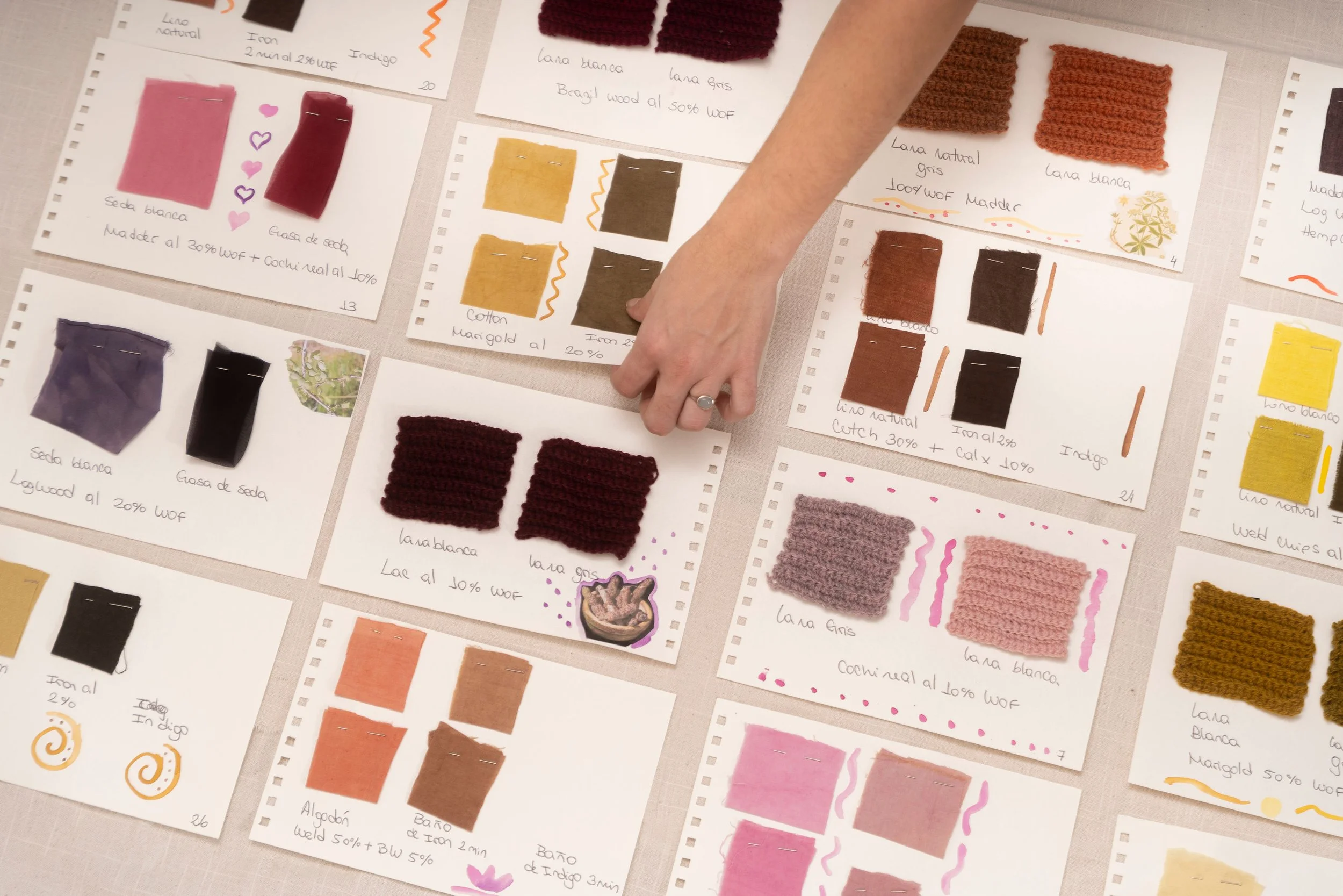 Flat lay of color swatches, yarn samples, and handwritten notes for a knitting or weaving project, with small fabric swatches pinned or attached to sheets of paper.