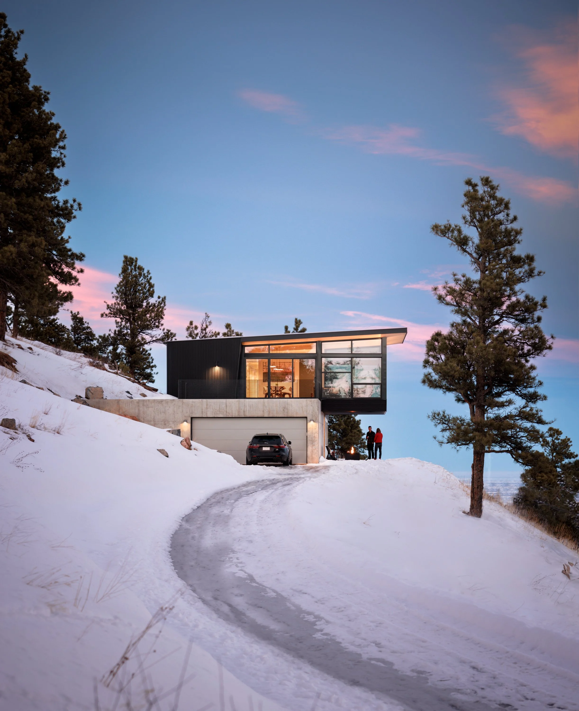 Modern house on a snowy hillside during sunset, with tall pine trees around and a car parked in front. Two people are standing near a campfire outside the house.