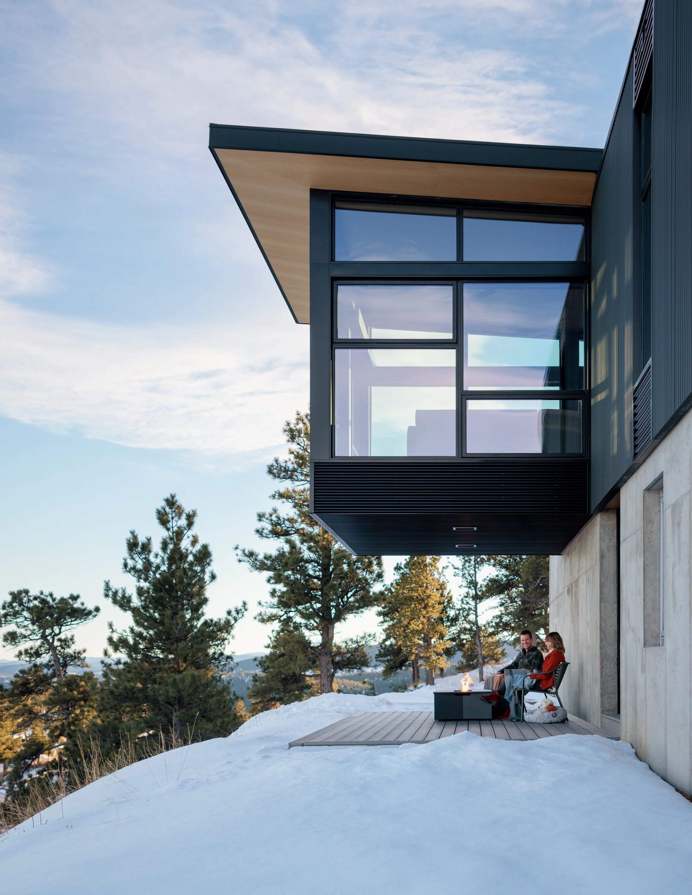Modern house with large glass windows and an overhanging roof, set in a snowy landscape with trees, two people sitting on chairs with a firepit between them, enjoying an outdoor winter scene.