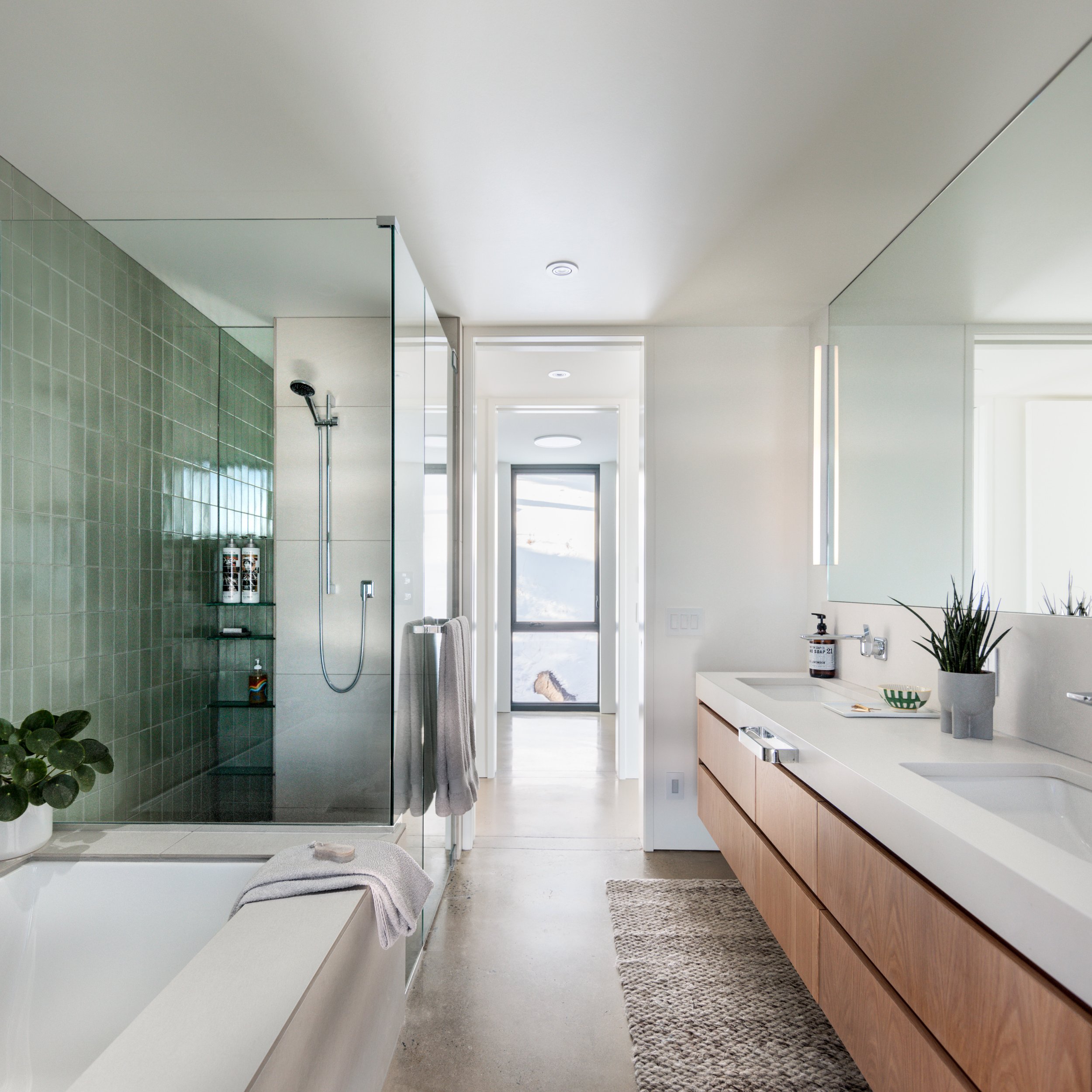 Modern bathroom with green tiled shower, glass partitions, white ceiling, large mirror, wooden cabinetry, and potted plant.