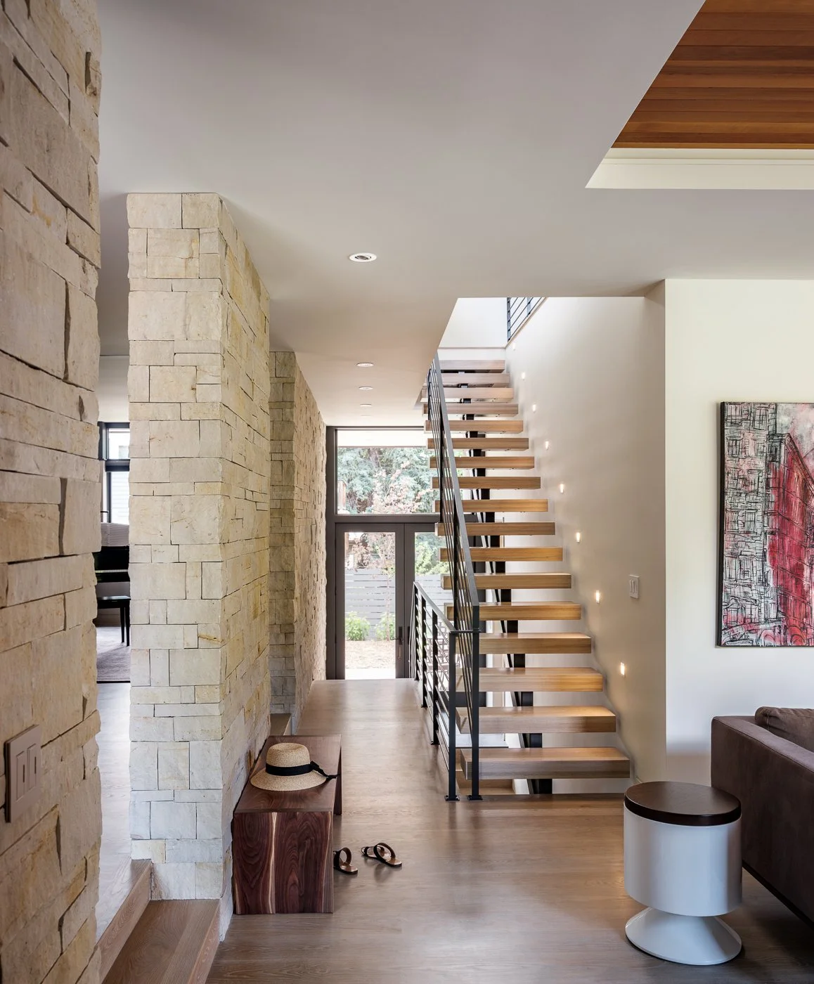 Modern interior entryway with stone walls, wooden staircase with metal railing, large window, and abstract art on the wall.