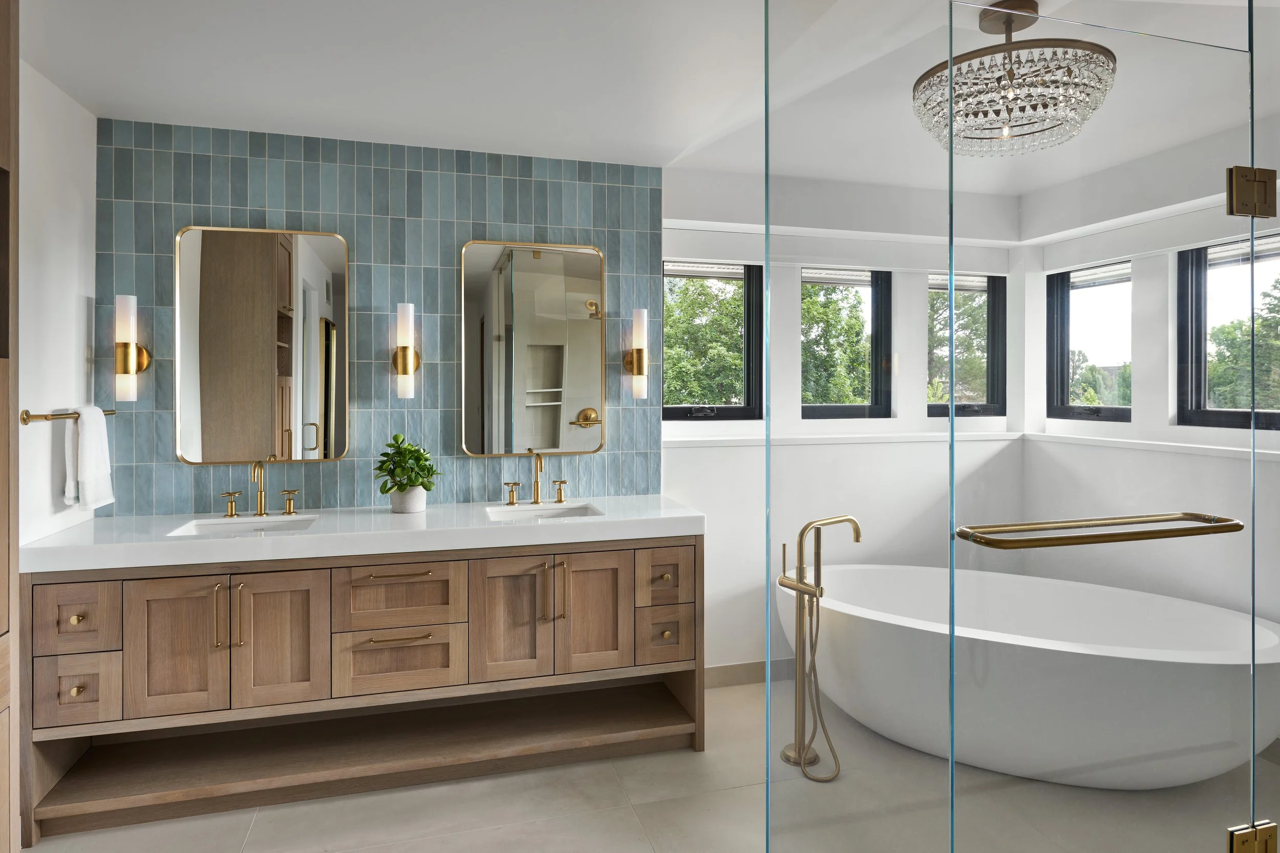 Modern bathroom with double vanity, two mirrors, gold fixtures, blue tiled wall, a plant, and a walk-in bathtub behind a glass partition.