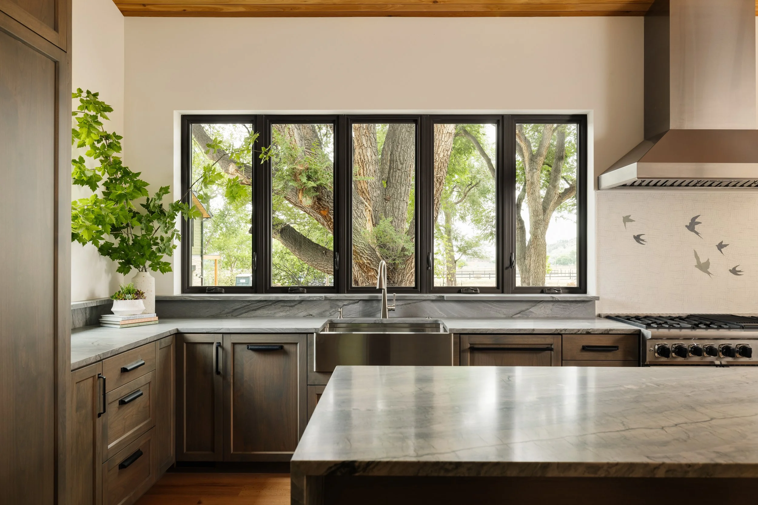 Kitchen with wood cabinetry, marble countertops, a large window showing trees outside, and a gray wall with bird decals.