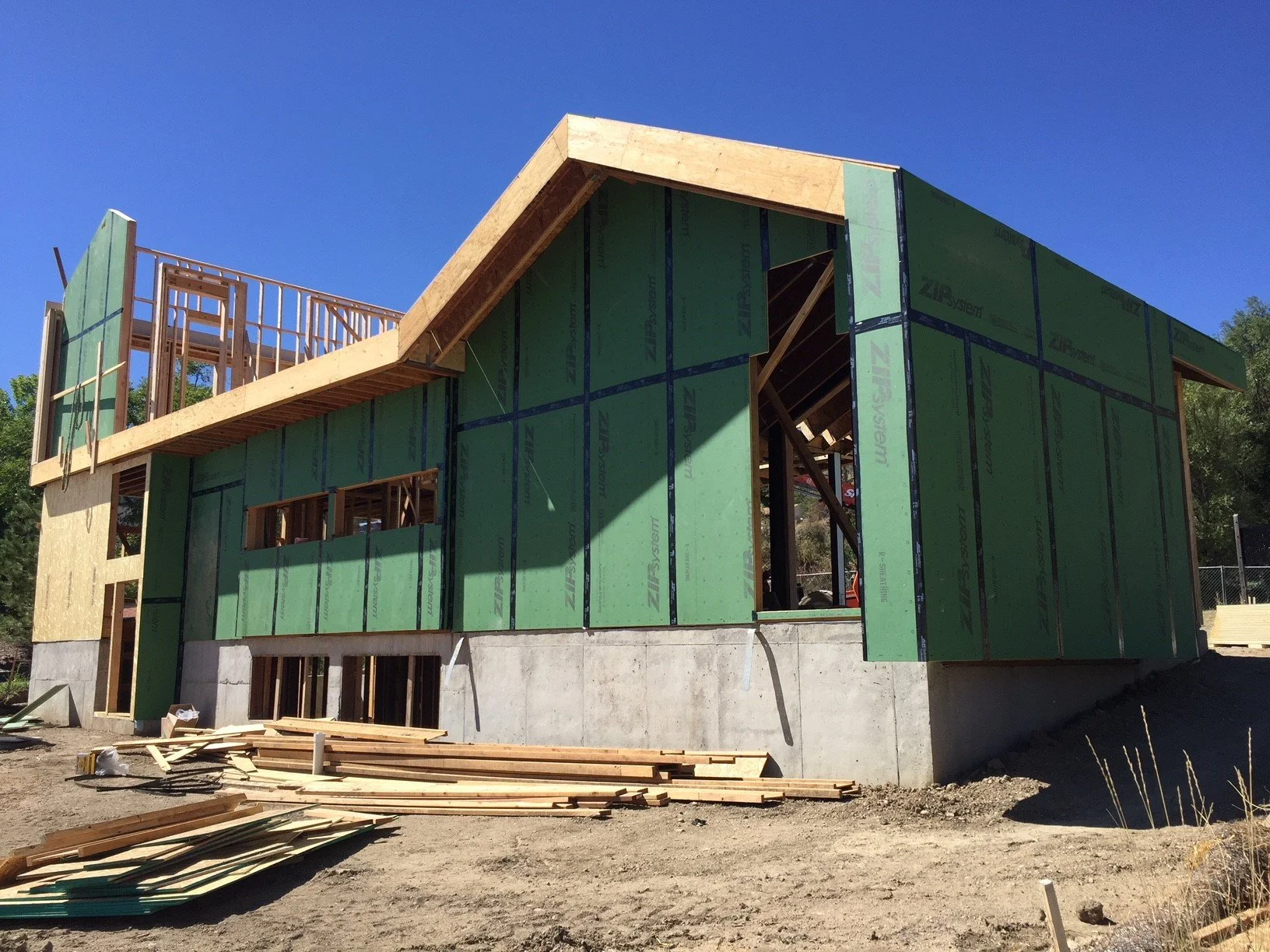 A house under construction with green sheathing panels and a concrete foundation, with wooden framing and a partially built roof.