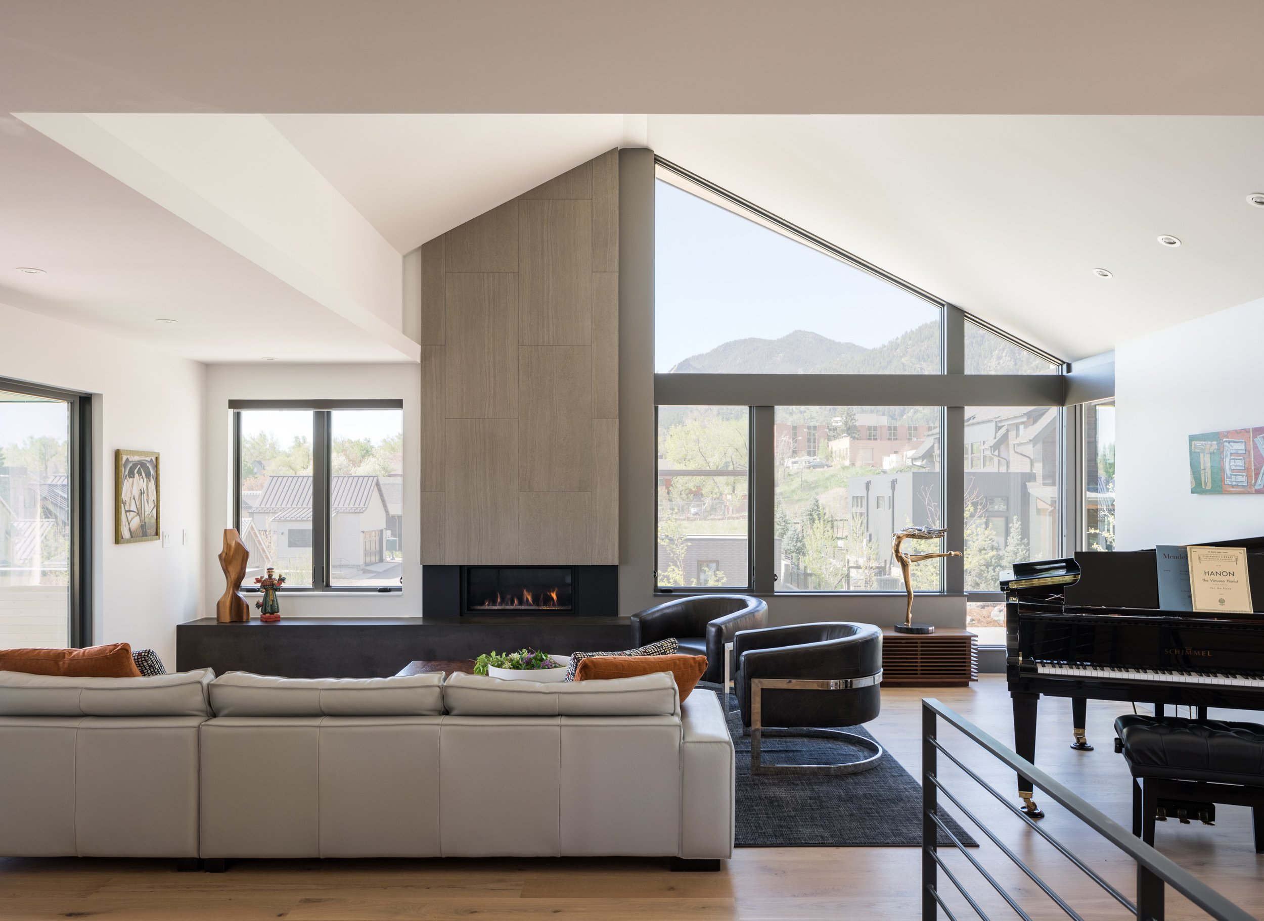 Modern living room with large windows, a fireplace, white sofa, black armchairs, and a black grand piano.