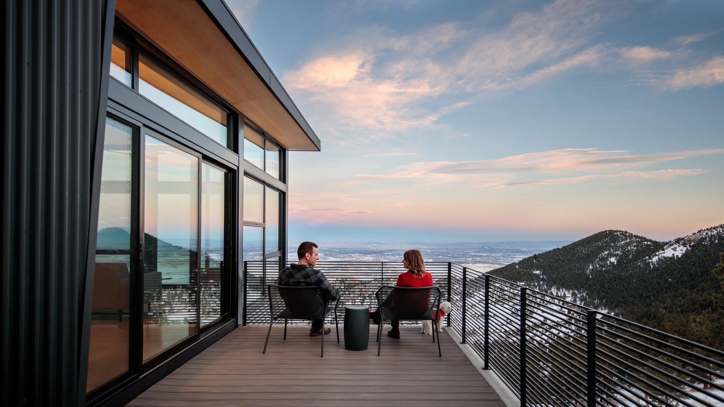 A man and woman sitting on a balcony outside a modern house with glass walls, overlooking a snowy mountainous landscape during sunset, with a small white dog nearby.