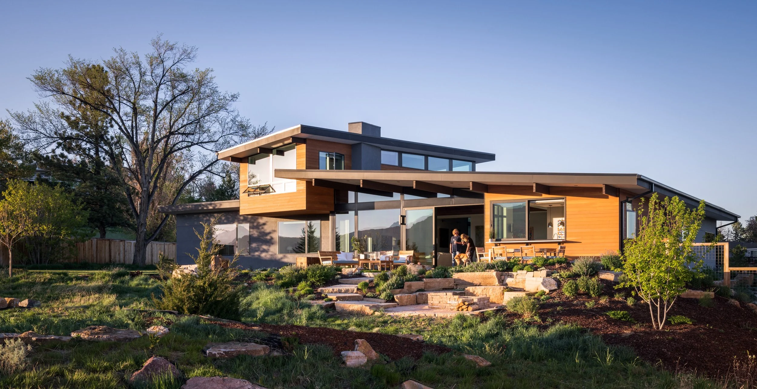 Modern house with large windows and a landscaped yard, with two people and a dog outside during sunset.