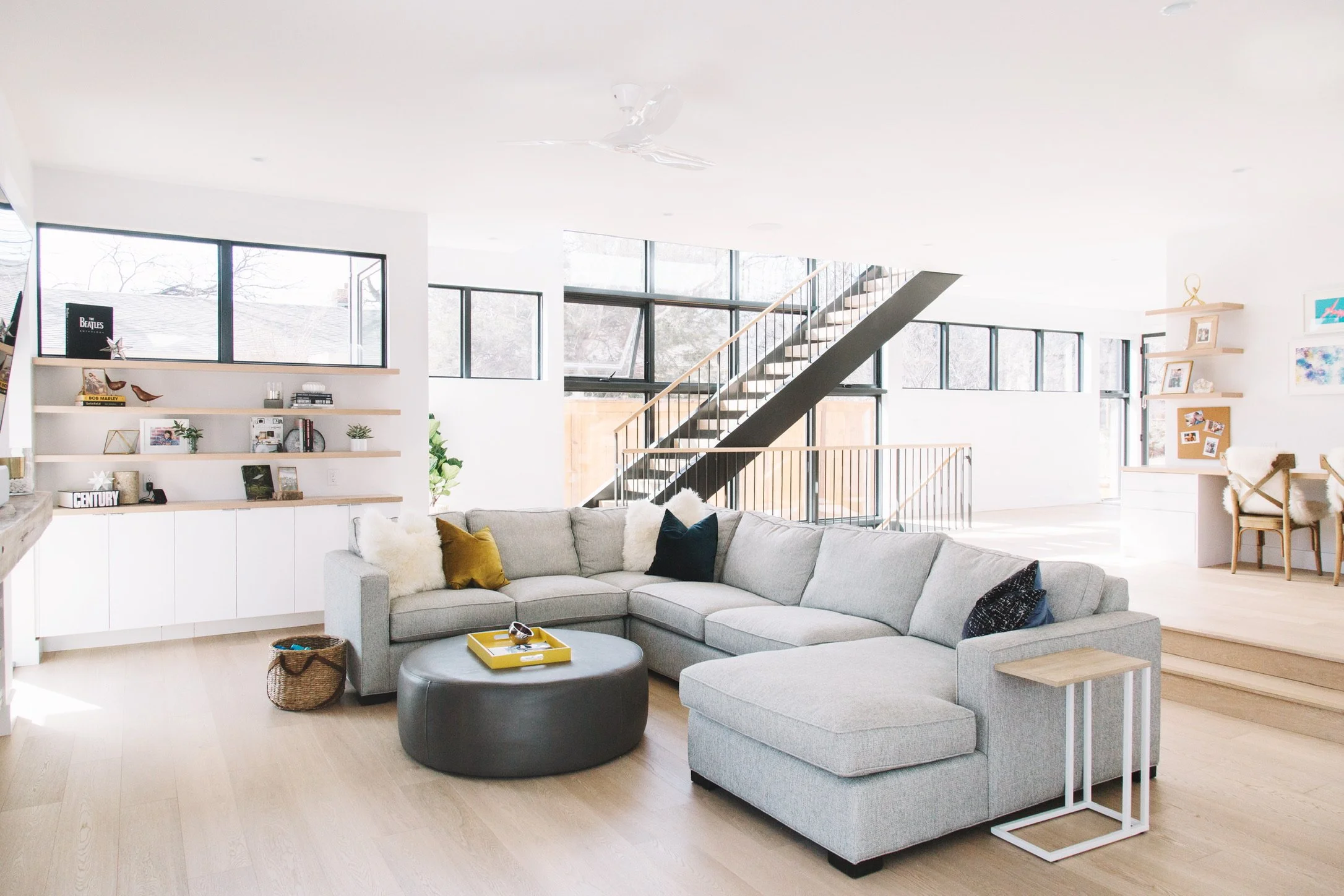 A spacious living room with large windows, a light gray sectional sofa, a round dark ottoman with a yellow tray, wooden shelves with decor, and stairs with black railing leading to the upper floor.
