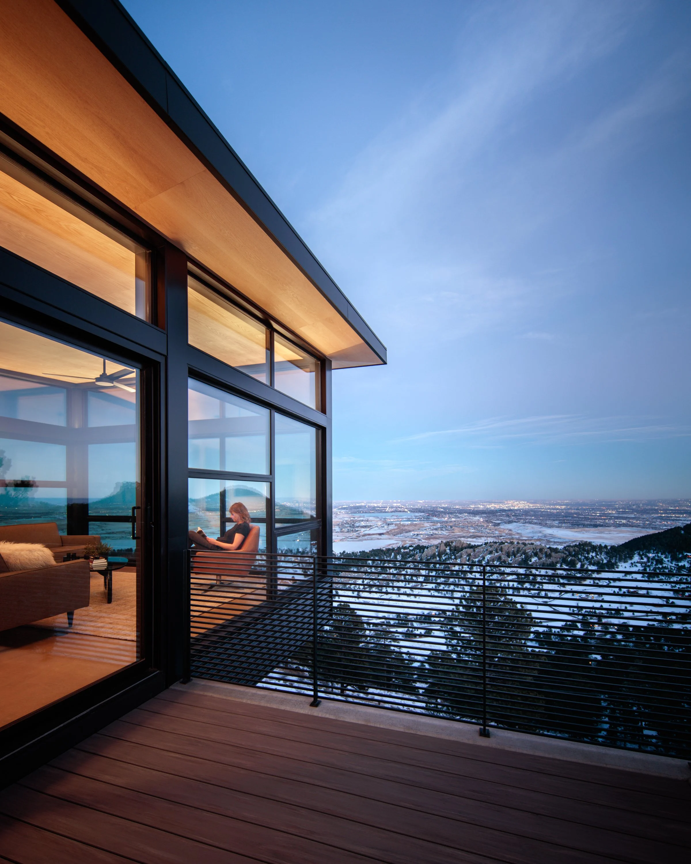 Modern house with large glass windows and wooden accents overlooking a snow-covered landscape and a cloudy blue sky.