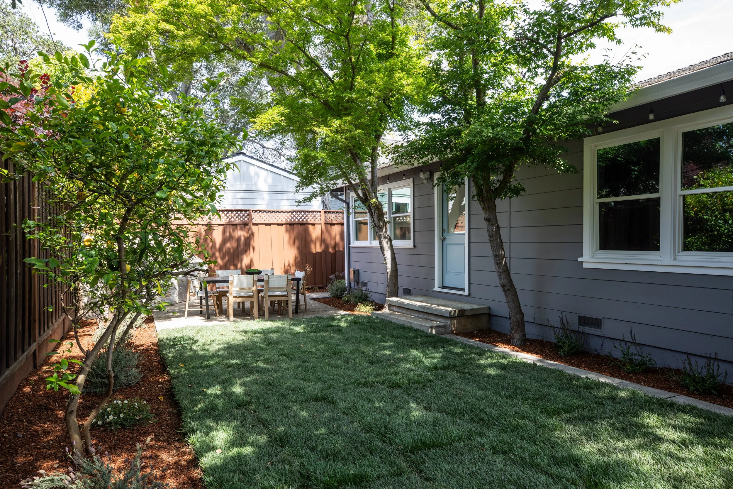 A backyard with a grassy lawn, surrounded by a wooden fence and mature trees providing shade. There is an outdoor dining area with a table and chairs near the house, which has gray siding and white-framed windows.