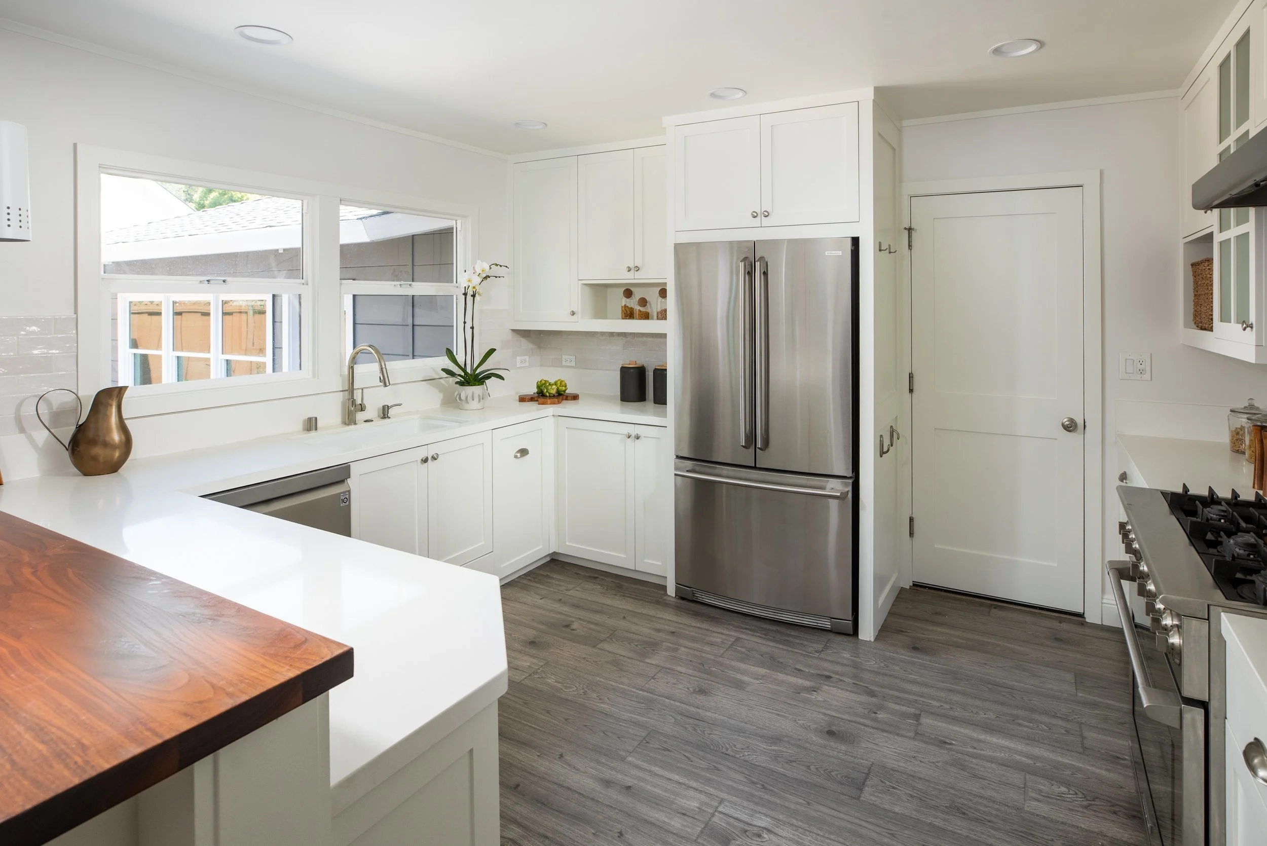 Bright modern kitchen with white cabinets, stainless steel refrigerator, wood flooring, large window above sink, and decorative items on counter.