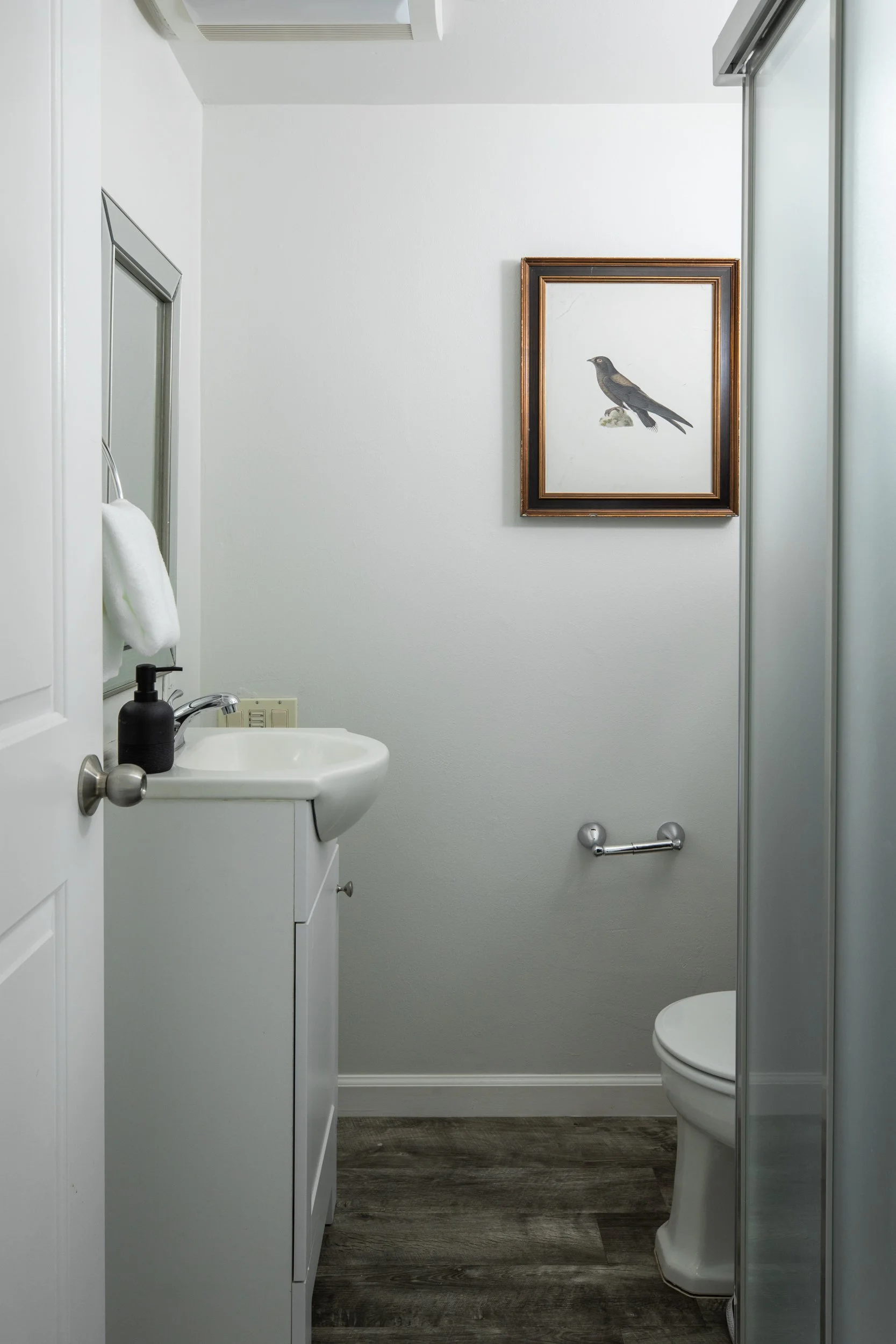 A small bathroom with white walls, a framed picture of a bird, a white sink with a black liquid soap dispenser, a towel hanging on a metallic towel ring, a toilet, and a mirrored shower enclosure.