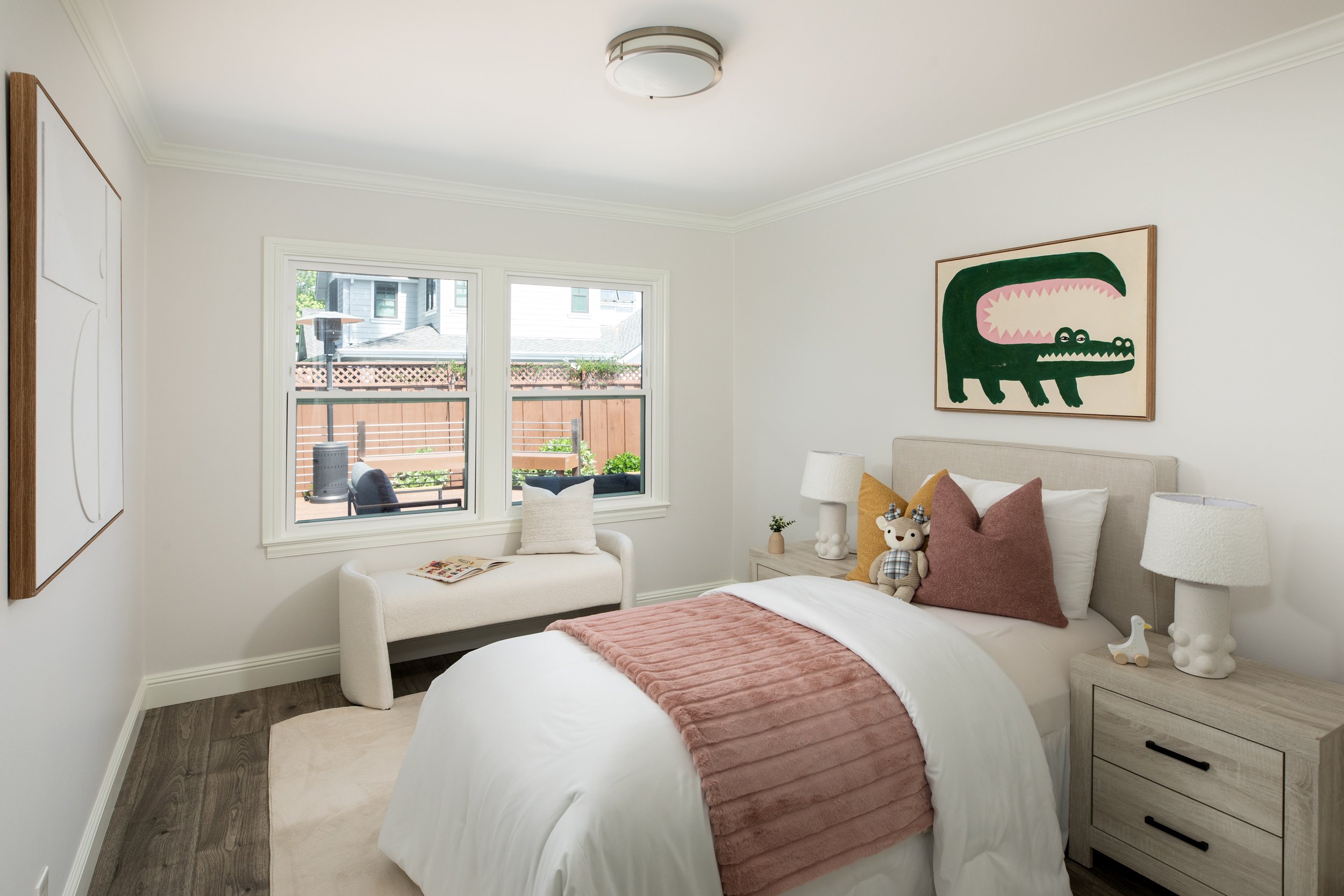 A cozy bedroom with a large bed covered with white bedding and a pink throw blanket. The bed has multiple pillows, including a brown one and a plaid stuffed animal. A white bedside table with decorative items, including a table lamp and a small plant