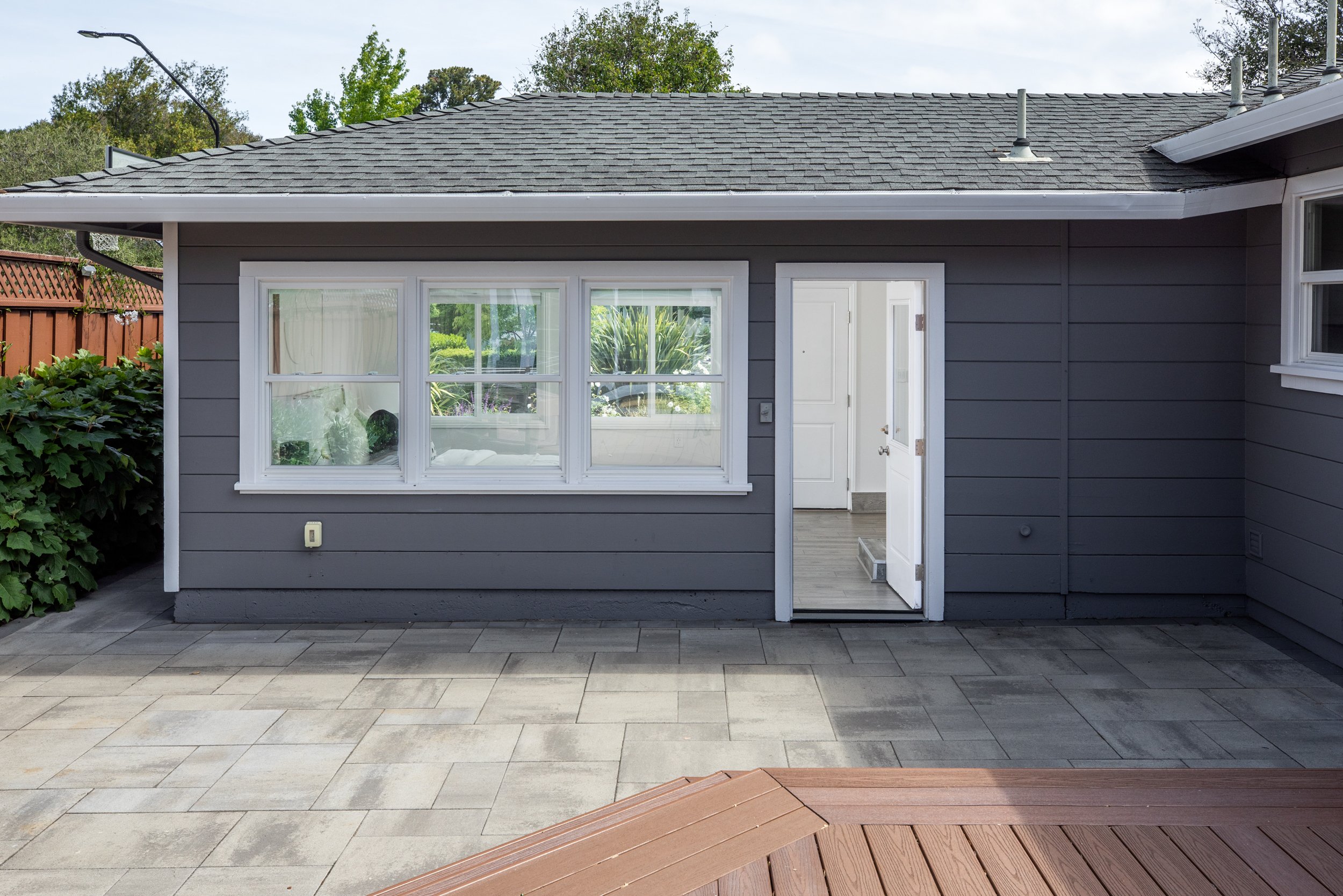 Backyard with gray house wall, large window, open door, tiled patio, wooden bench, and garden plants.