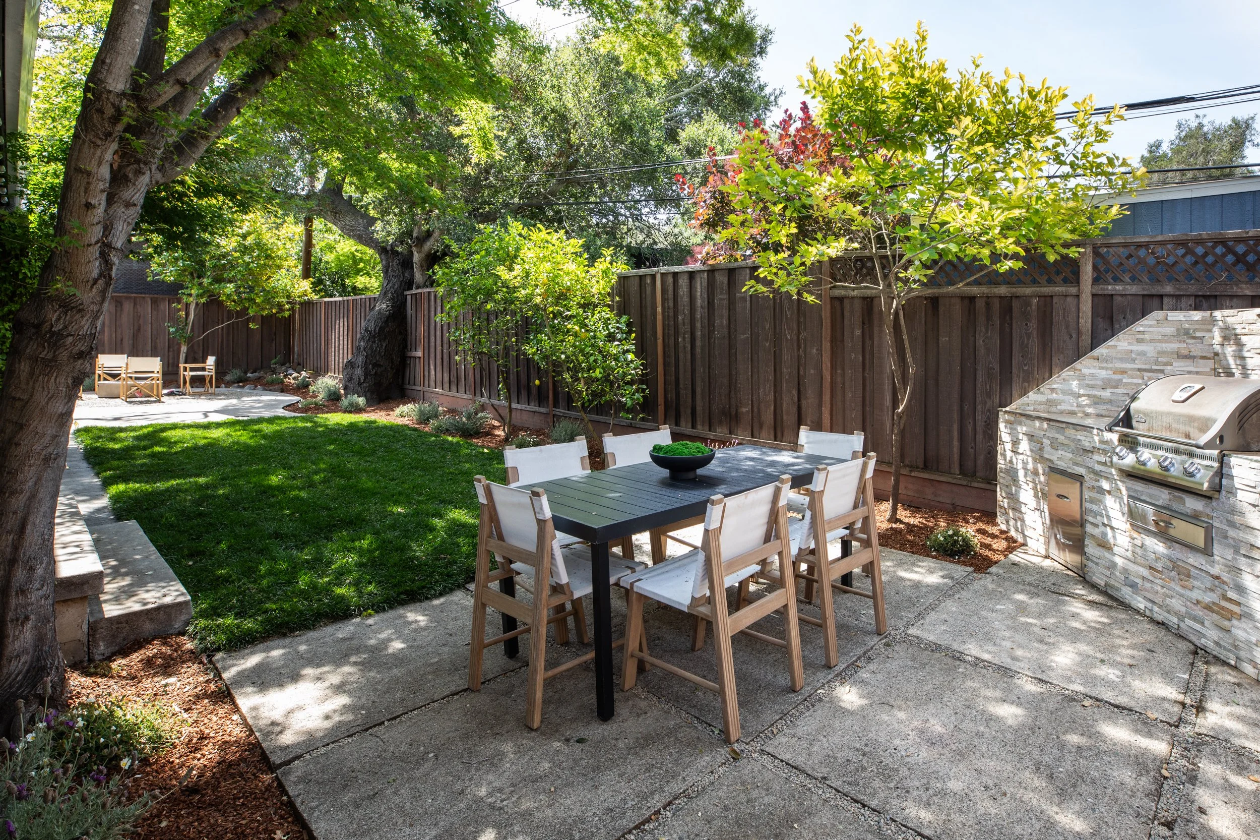 A backyard patio area with a wooden dining table and chairs, surrounded by landscaped garden beds with various trees, plants, and shrubs. There is an outdoor grill built into a stone structure on the right side. The yard is enclosed by a wooden fence