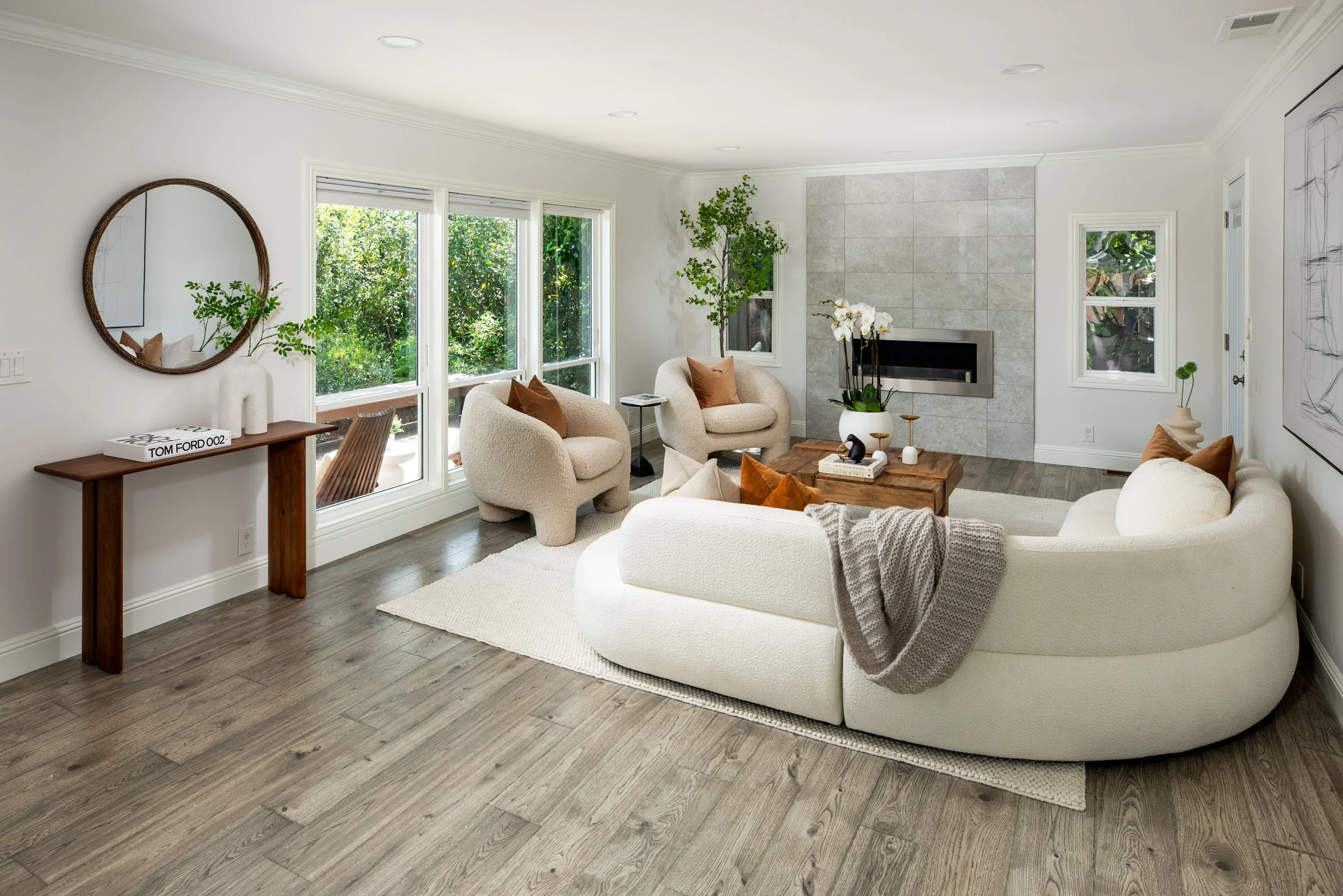 A bright living room with large windows, white walls, a white sectional sofa with brown pillows, a circular mirror on the wall, a wooden console table with a potted plant, and a fireplace with a decorative orchid, all on a wood floor.