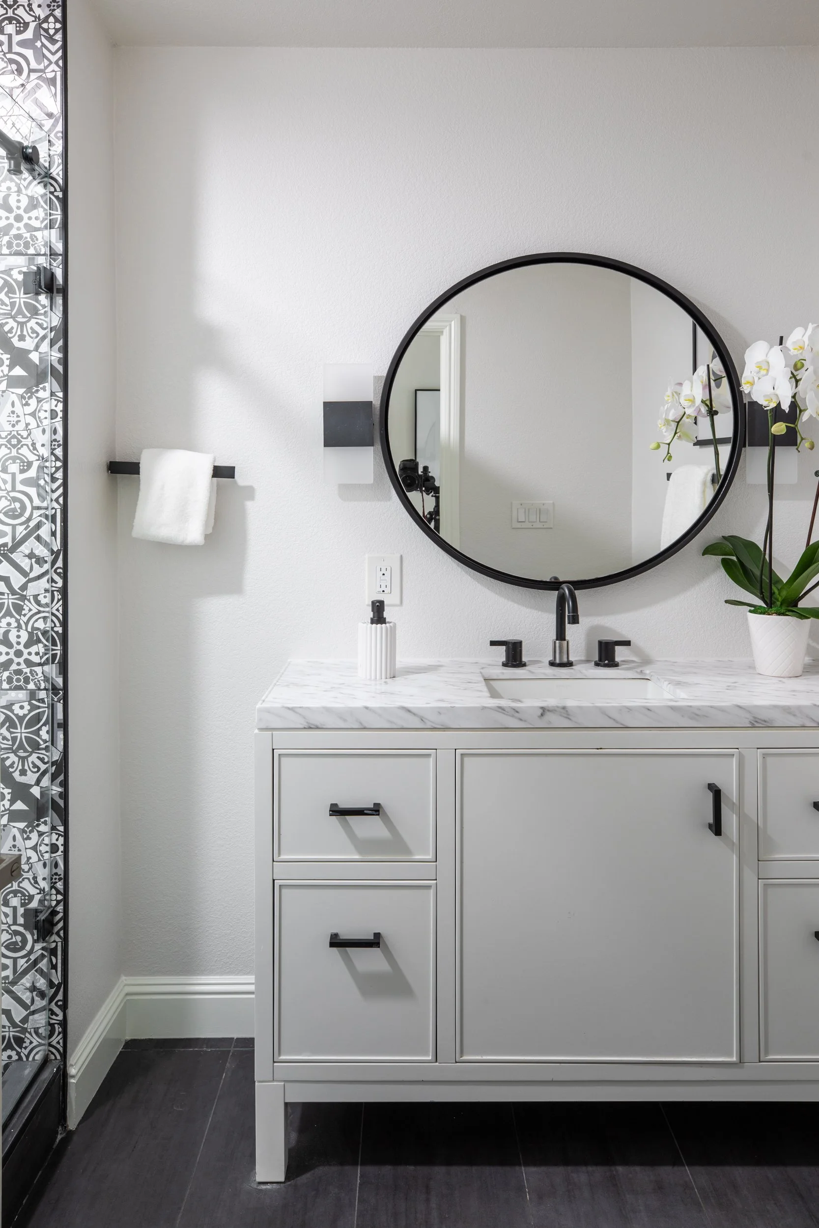 A bathroom with a white vanity cabinet with three drawers, a marble countertop, and a black faucet. Above the sink is a round mirror, and a white orchid plant is on the right side of the countertop. There is a towel on a black towel bar to the left o