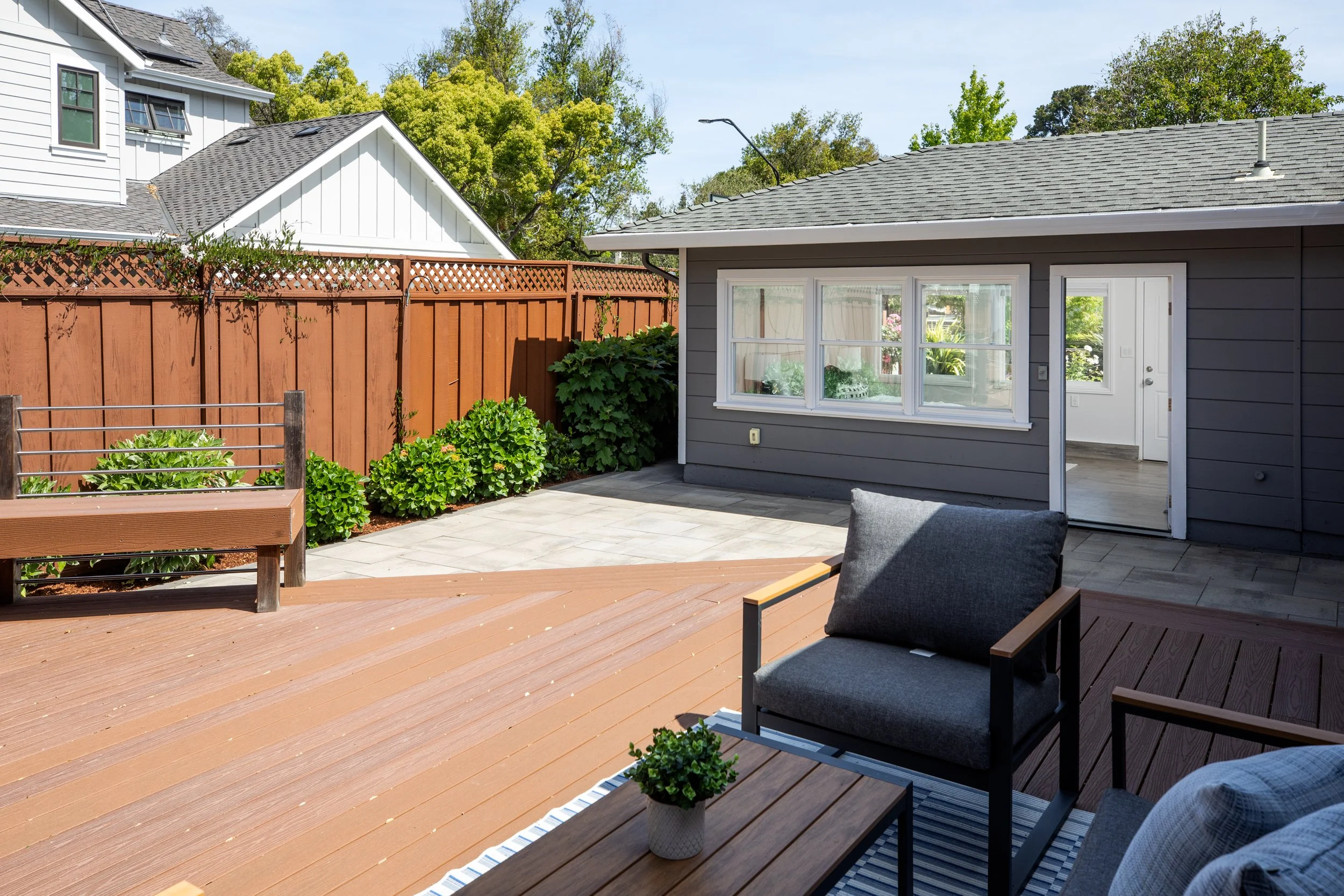 A backyard with a wooden deck, a gray house with large windows, a wooden fence, and green plants. There are outdoor chairs with cushions and a small side table with a potted plant.
