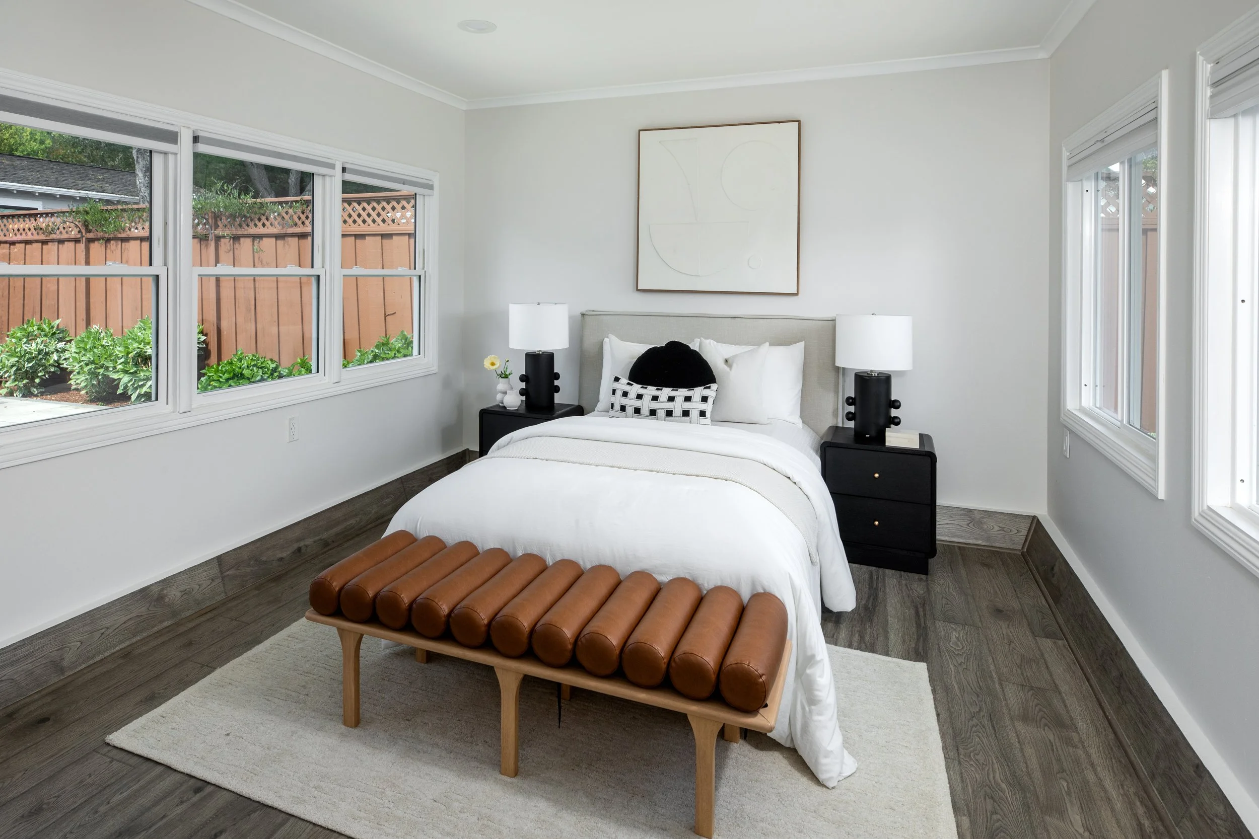 A bedroom with white walls, large windows, a bed with a white comforter, black nightstands with white lamps, a brown leather bench, and minimalist artwork.