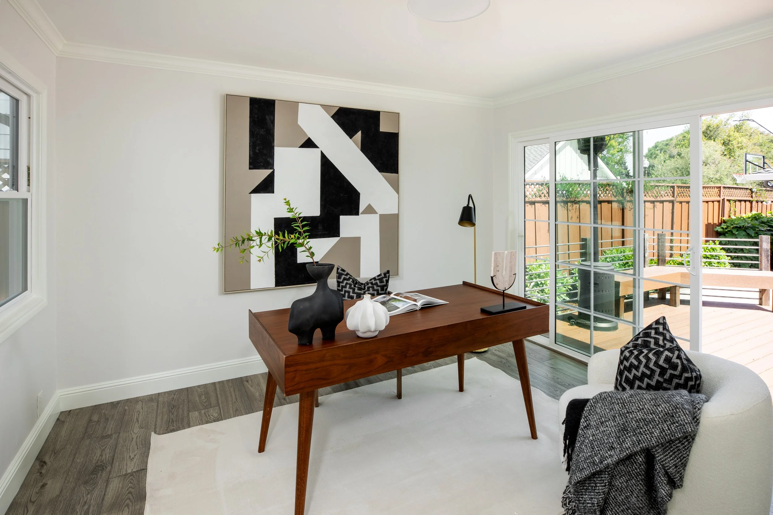 The image displays a bright living room with white walls, a large window with sliding glass doors showing a backyard with a wooden deck, greenery, and a basketball hoop. There is a wooden table with decorative items, including a black vase with a gre