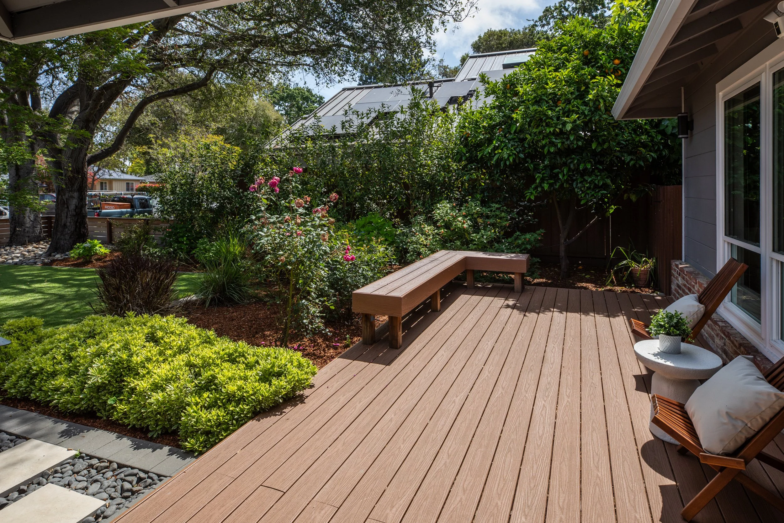 A backyard patio with a wooden deck, surrounded by lush greenery and flowering bushes, adjacent to a gray house with large windows, and trees in the background.