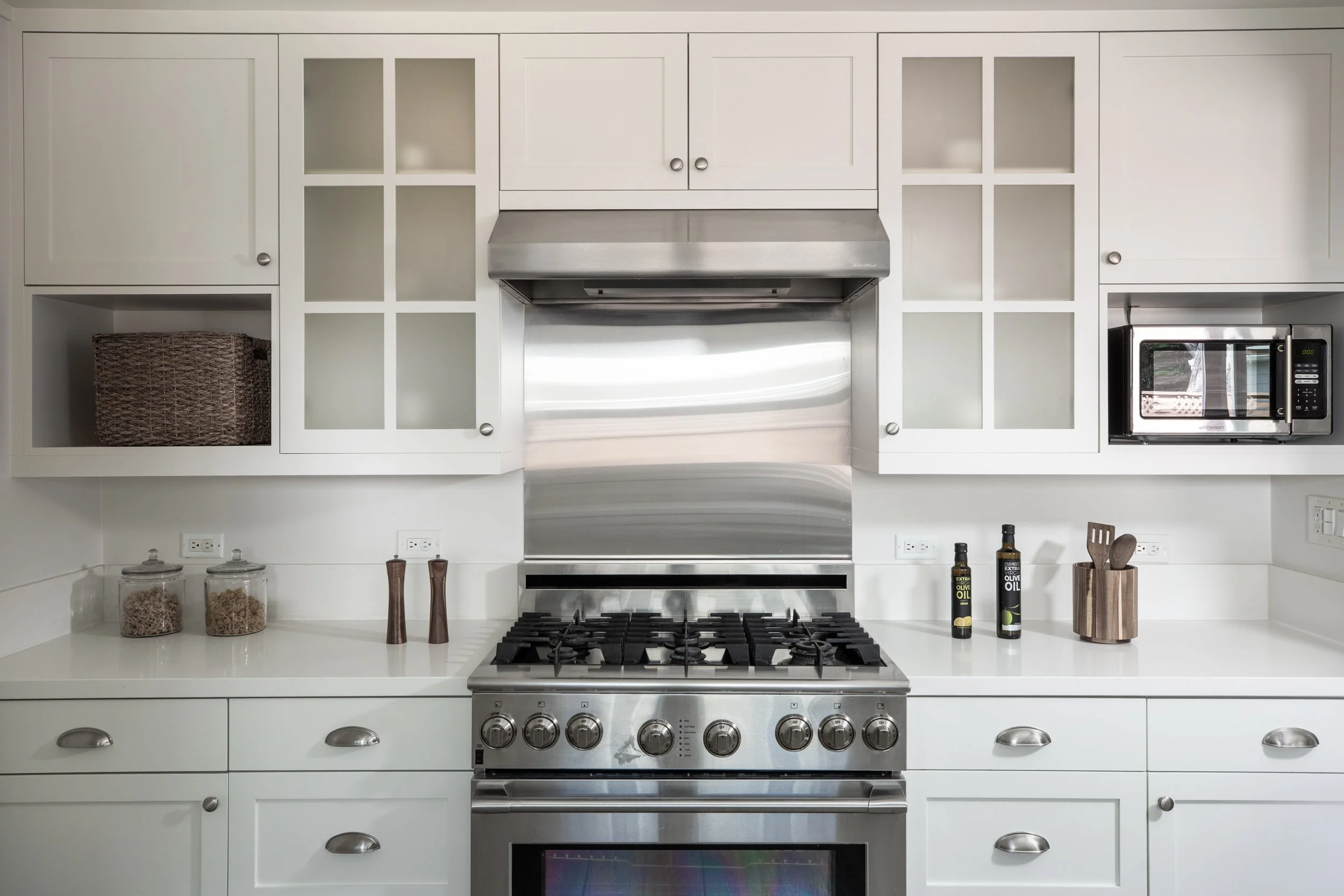Kitchen with white cabinets, stainless steel range, microwave, and countertops. Items include glass jars with dry pasta, olive oil bottles, and wooden utensils.