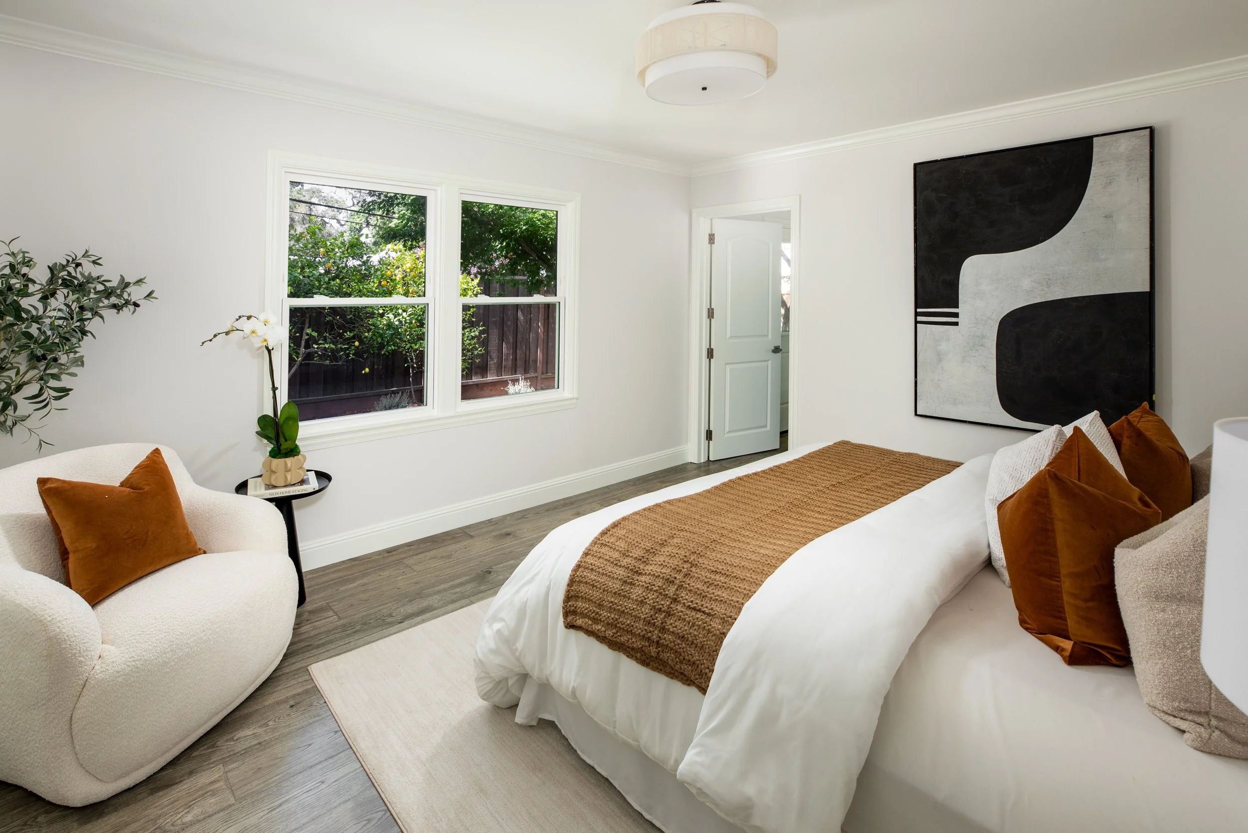 A bedroom with white walls, a bed with a brown blanket and orange pillows, a cream-colored armchair with orange pillows, a small black side table with a book and plant, a large abstract black and white painting on the wall, a window showing greenery 