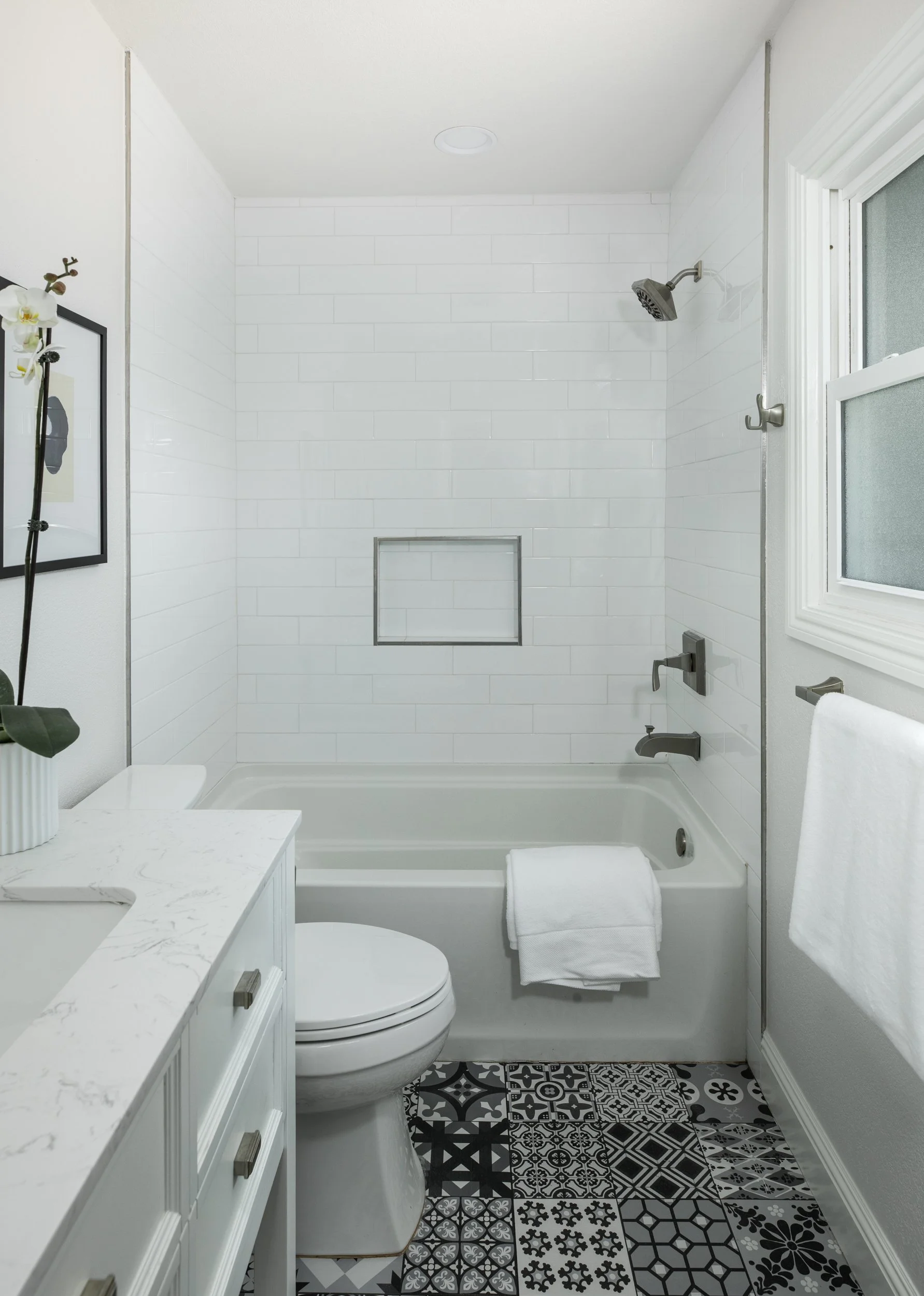 Modern bathroom with white tile shower, white vanity with marble countertop, and patterned black and white floor tiles. A toilet with a rolled towel on top is seen next to the vanity, and a window with frosted glass is on the right. There is a small 