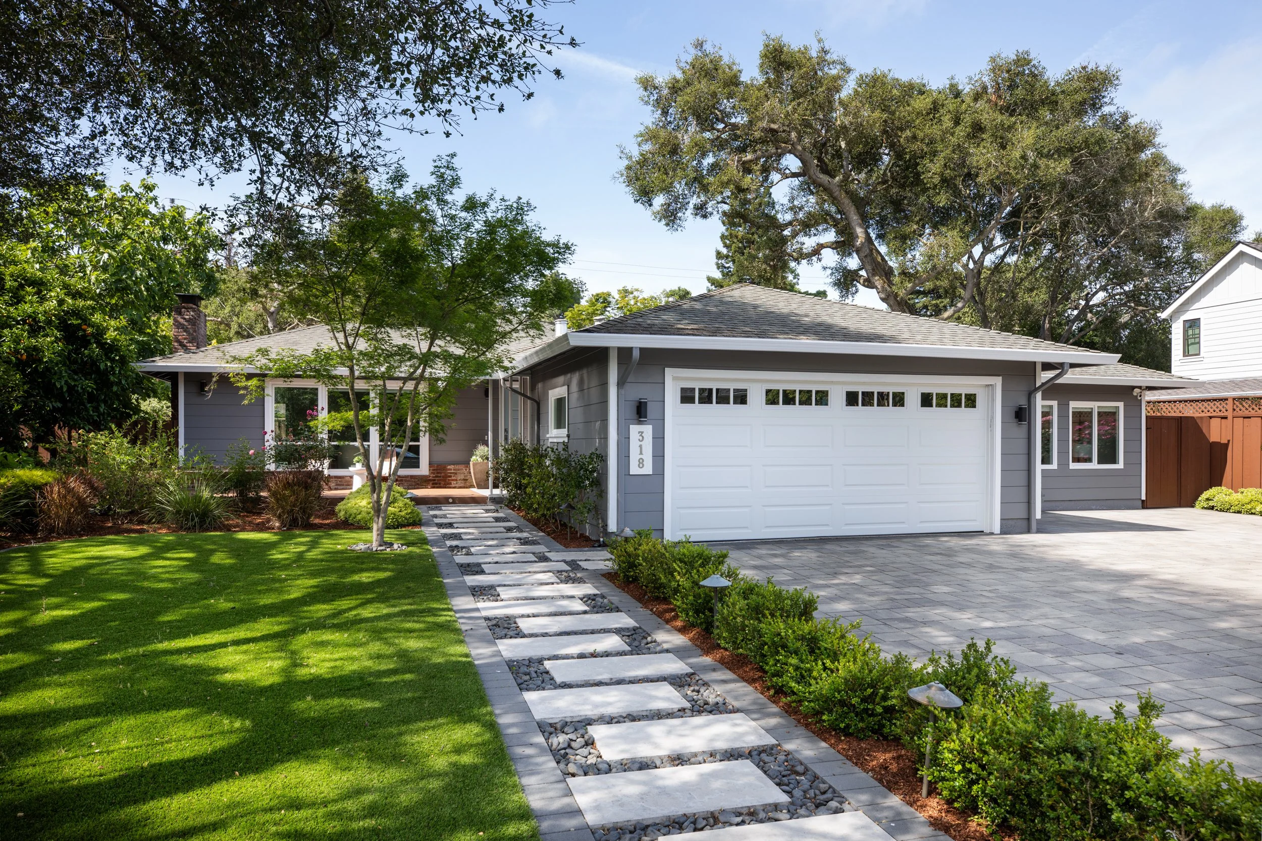 A modern residential house with gray siding, a white garage door, and a landscaped front yard featuring a stone pathway, green lawn, and trees under a blue sky.