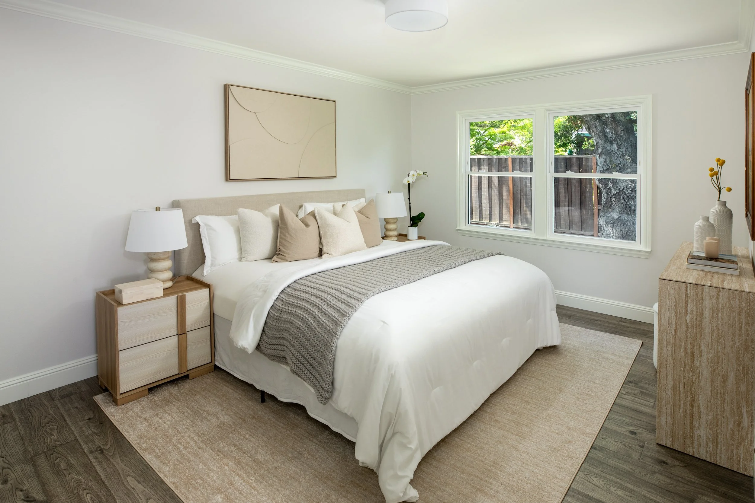 A bedroom with a neatly made bed, beige and white pillows, and a gray blanket. There are two wooden nightstands with white lamps on either side of the bed. A large window shows trees outside. Decorative vases and a book are on a dresser to the right.