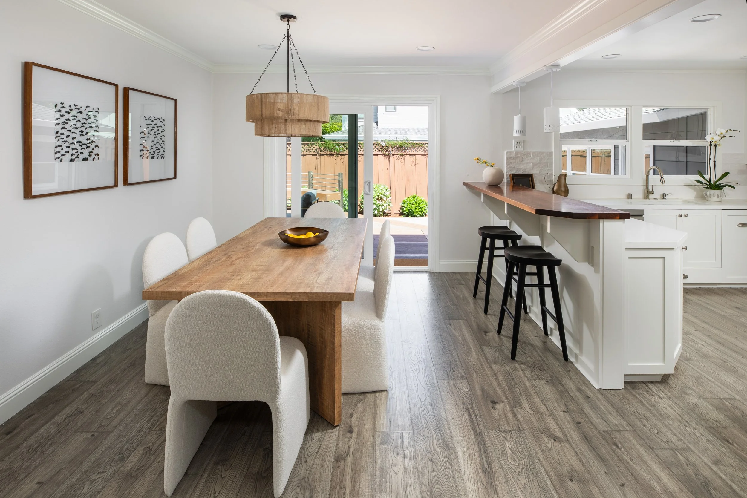 Dining area with a wooden table surrounded by six upholstered chairs, a hanging light fixture, and artwork on the wall. The room opens to an outdoor patio through a sliding glass door.