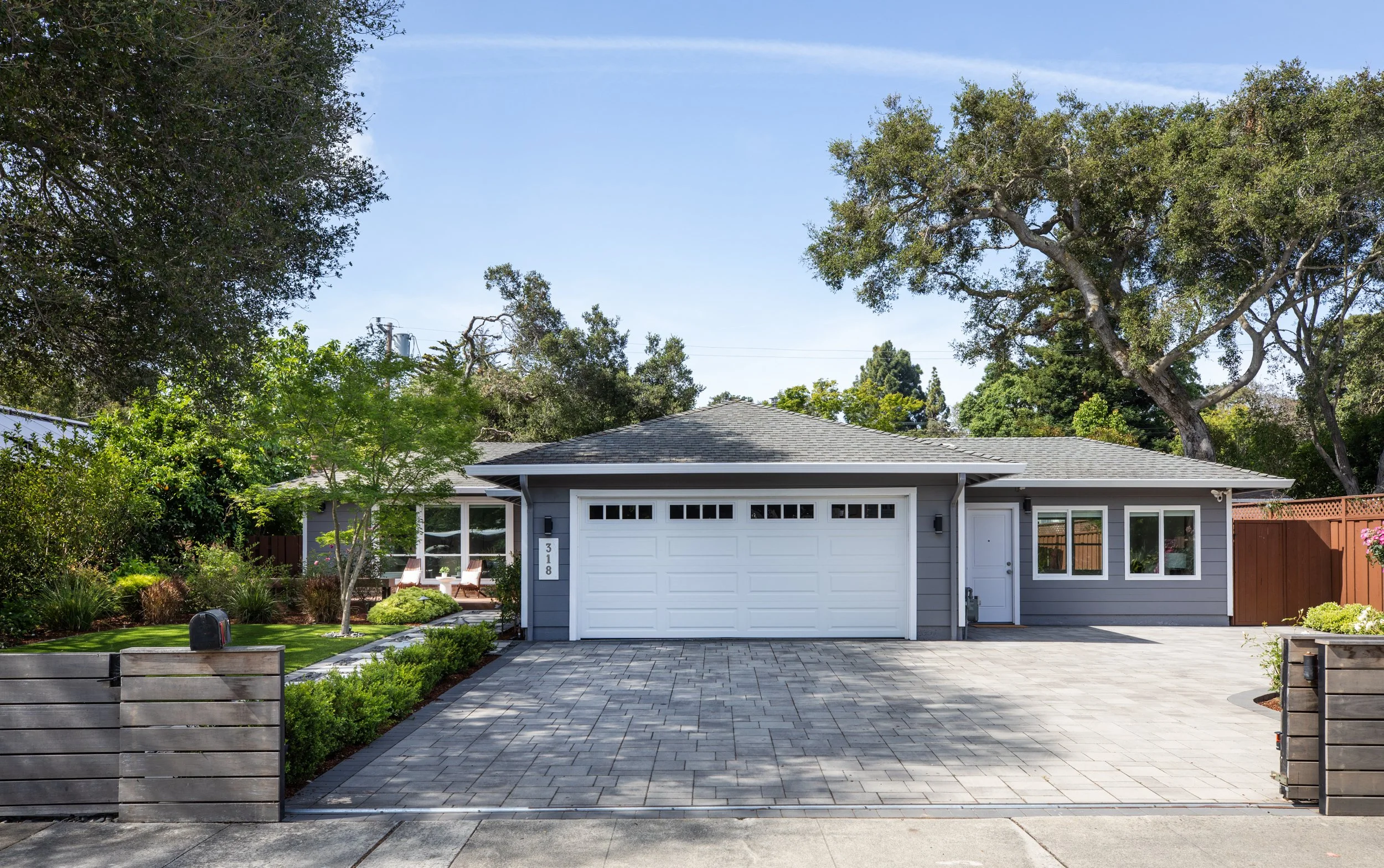 Front view of a gray house with a white garage door, surrounded by green trees and a well-maintained garden, with a paved driveway and a wooden fence on the sides.