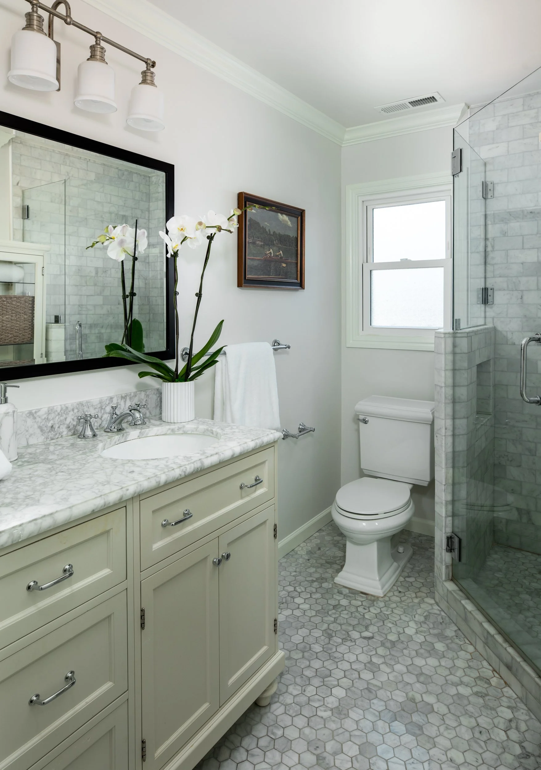 A bathroom with a white and gray marble vanity with a sink, mirror, and white orchids. A toilet is next to the vanity, and a shower with a glass door is in the corner. There is a window above the toilet, and a framed painting hangs on the wall.