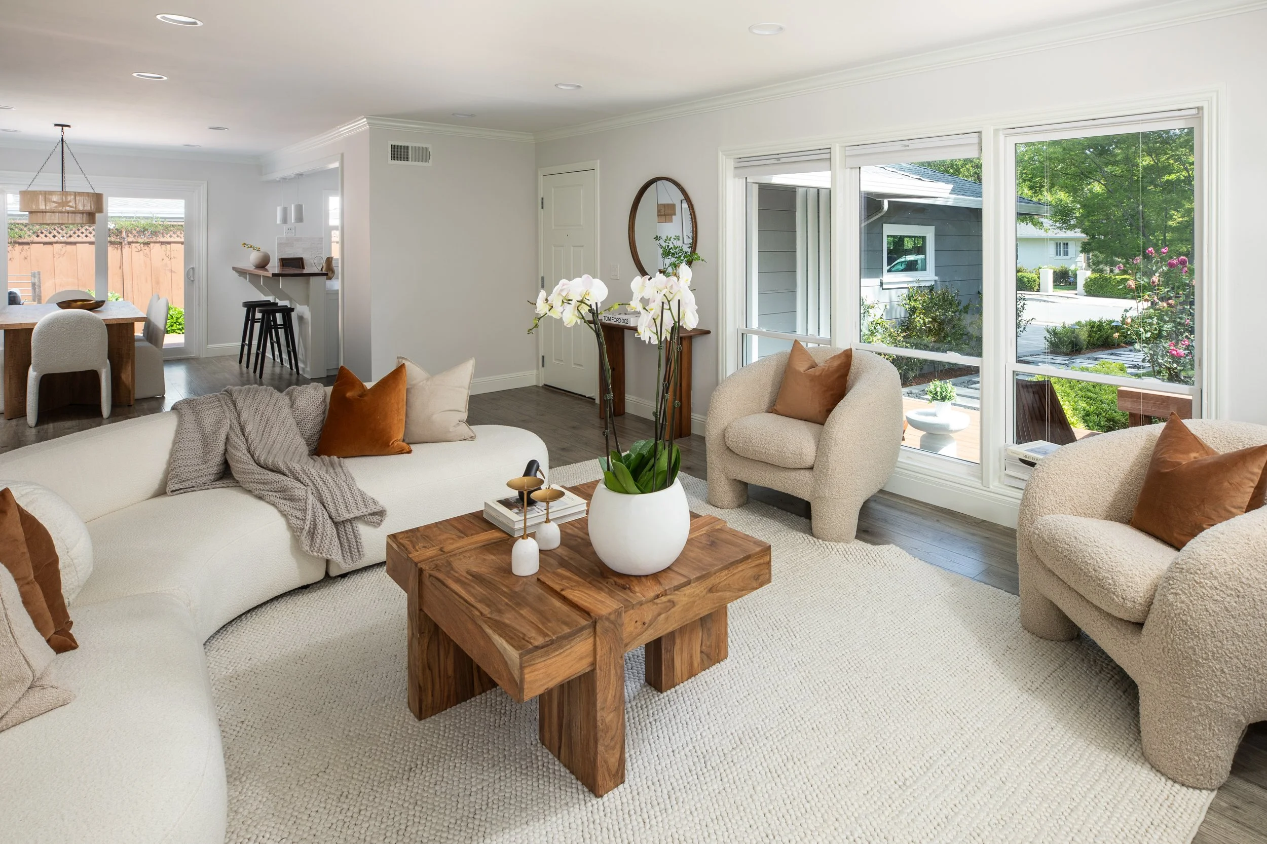 Bright living room with large windows, beige and white furniture, and a wooden coffee table with orchids and decorative items. Open dining area with a wooden table and white chairs, view of a backyard outside.
