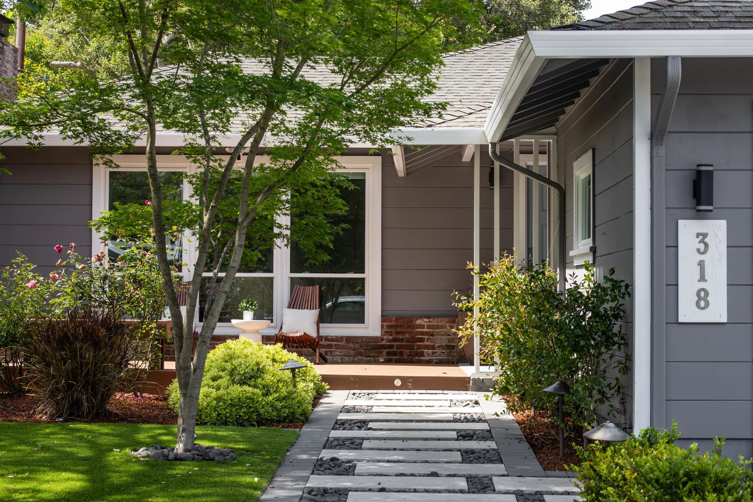 Front yard of a house with a gray exterior, large window, and a path made of stepping stones bordered by small plants and rocks. There is a tree with green leaves, shrubs, and a small sitting area with chairs and a side table in front of the house.