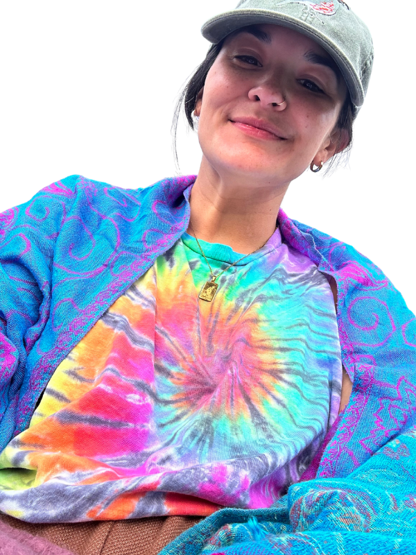 A white person wearing a colorful tie-dye shirt, a purple patterned jacket, a gray cap, and a gold necklace with a pendant, smiling at the camera.