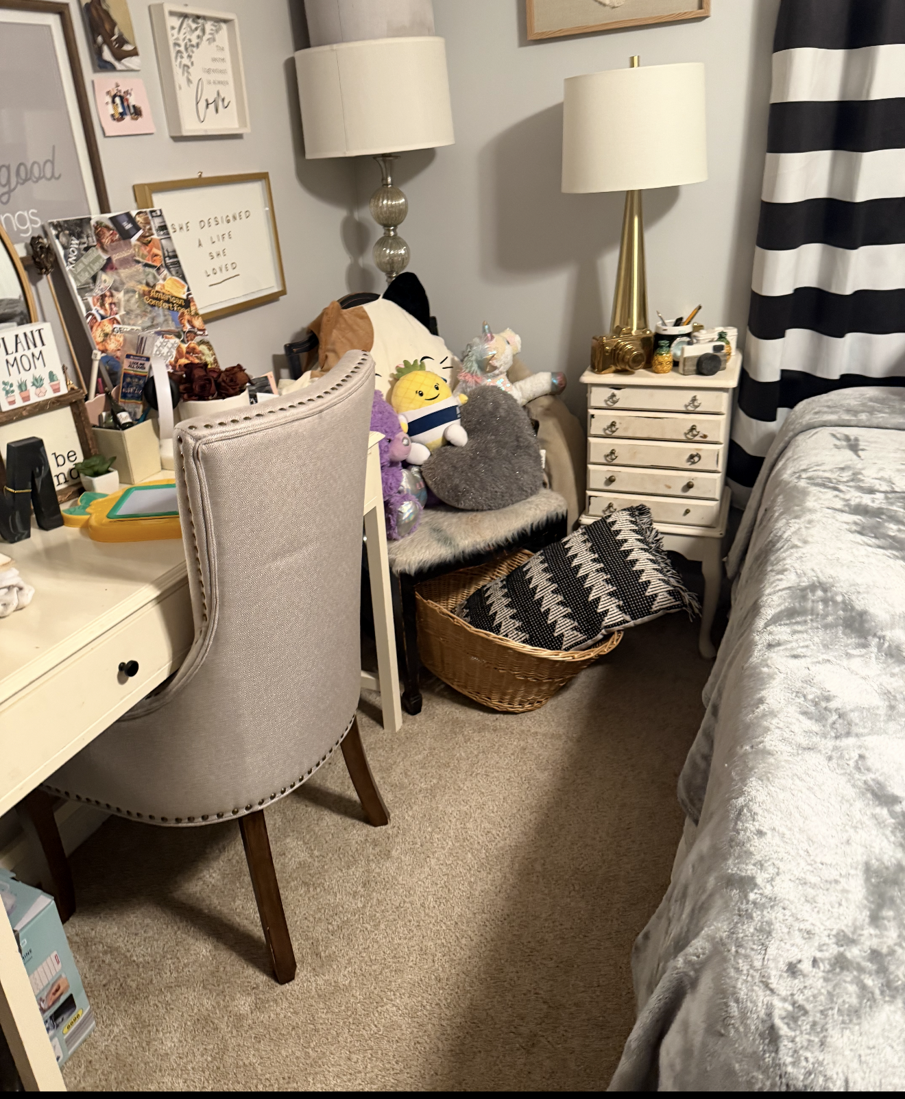 A bedroom corner with a beige desk, a beige upholstered chair, a white nightstand with gold knobs, two white lamps, a bed with a gray floral cover, a basket with a black and white blanket, and stuffed toys on a black chair. Wall decorations include framed quotes and artwork, and a crumpled hoodie is on the headboard.