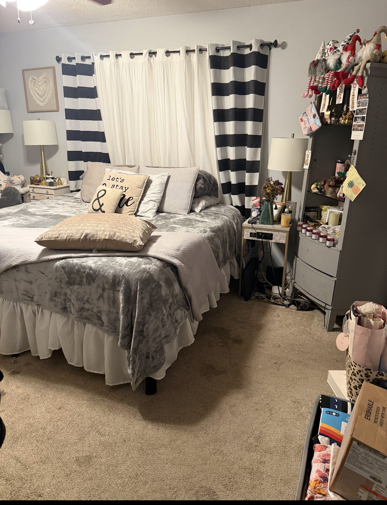 A cozy bedroom with a bed covered in a velvet grey blanket and multiple pillows, including one with the phrase "Let's stay & love." There are two nightstands with lamps beside the bed, and a window with white curtains and navy blue-striped drapes. To the right, a tall grey shelf holds Christmas stockings, small gifts, and decorative items. Various items are on the floor and surfaces, including a bouquet of dried flowers, candles, and miscellaneous objects.