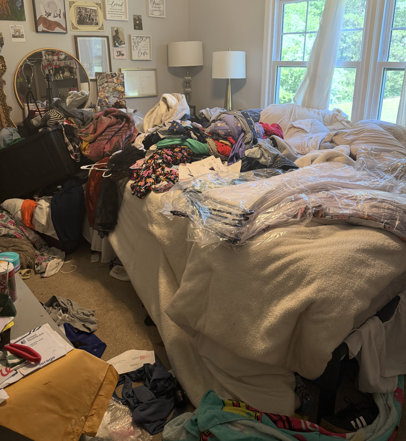 A cluttered bedroom with a bed covered in clothes and plastic bags, a wall decorated with framed pictures and artwork, and a window with white curtains letting in natural light.