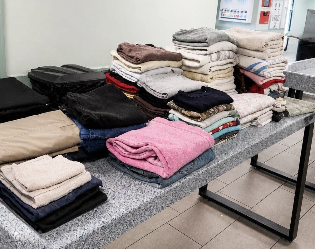 Stacks of folded clothes, including towels, shirts, and pants, on a counter in a laundromat.