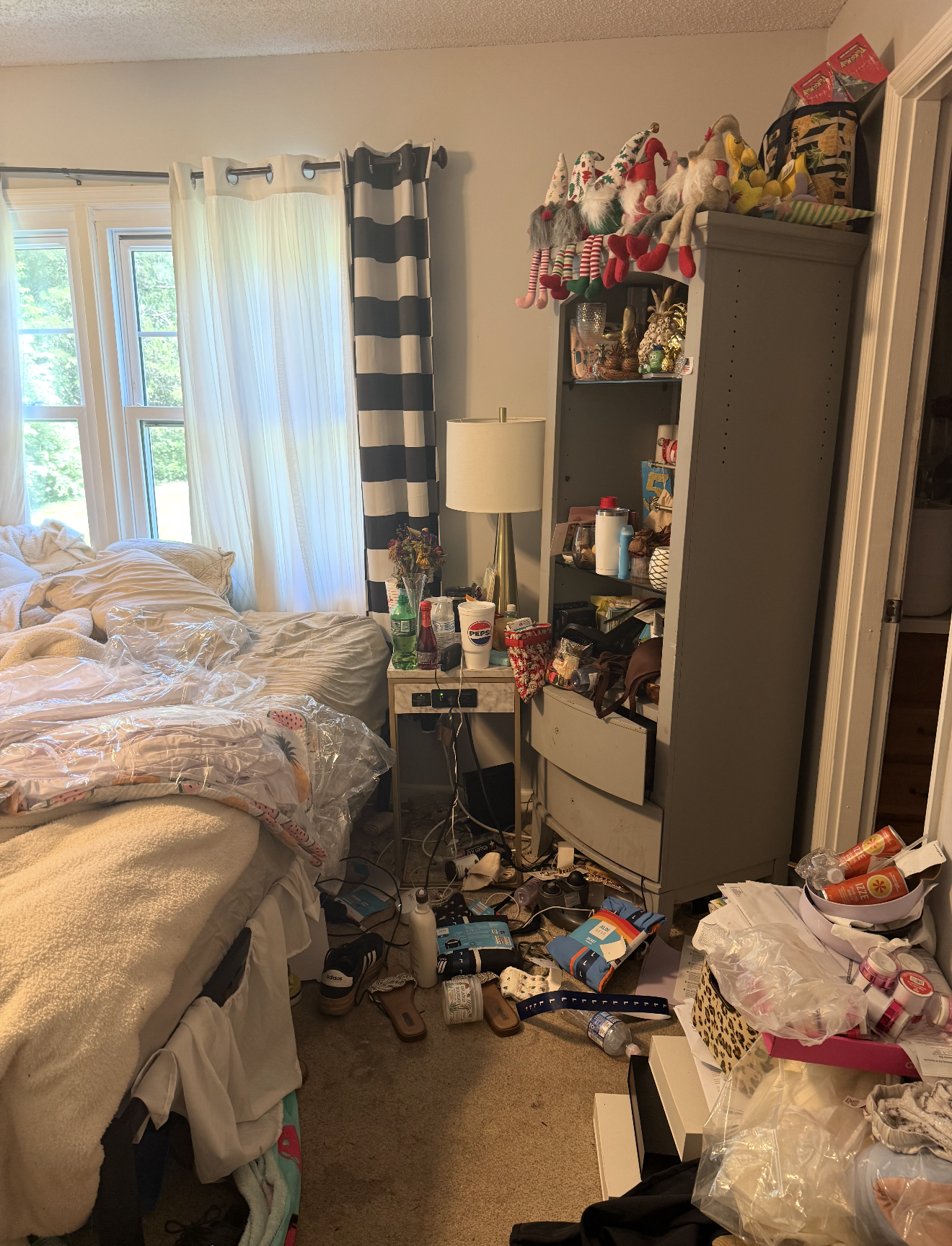 A cluttered bedroom with a bed, nightstand, and shelves, containing various items like drinks, snacks, Christmas decorations, and miscellaneous clutter.