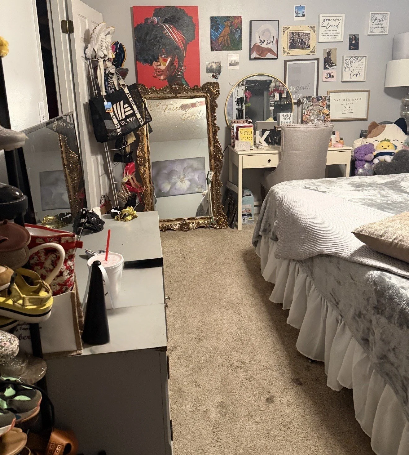 A cluttered bedroom with a bed, a white desk with various items on it, a large decorated mirror, and wall art including a colorful portrait of a woman with natural hair. The wall has multiple framed pictures and quotes.