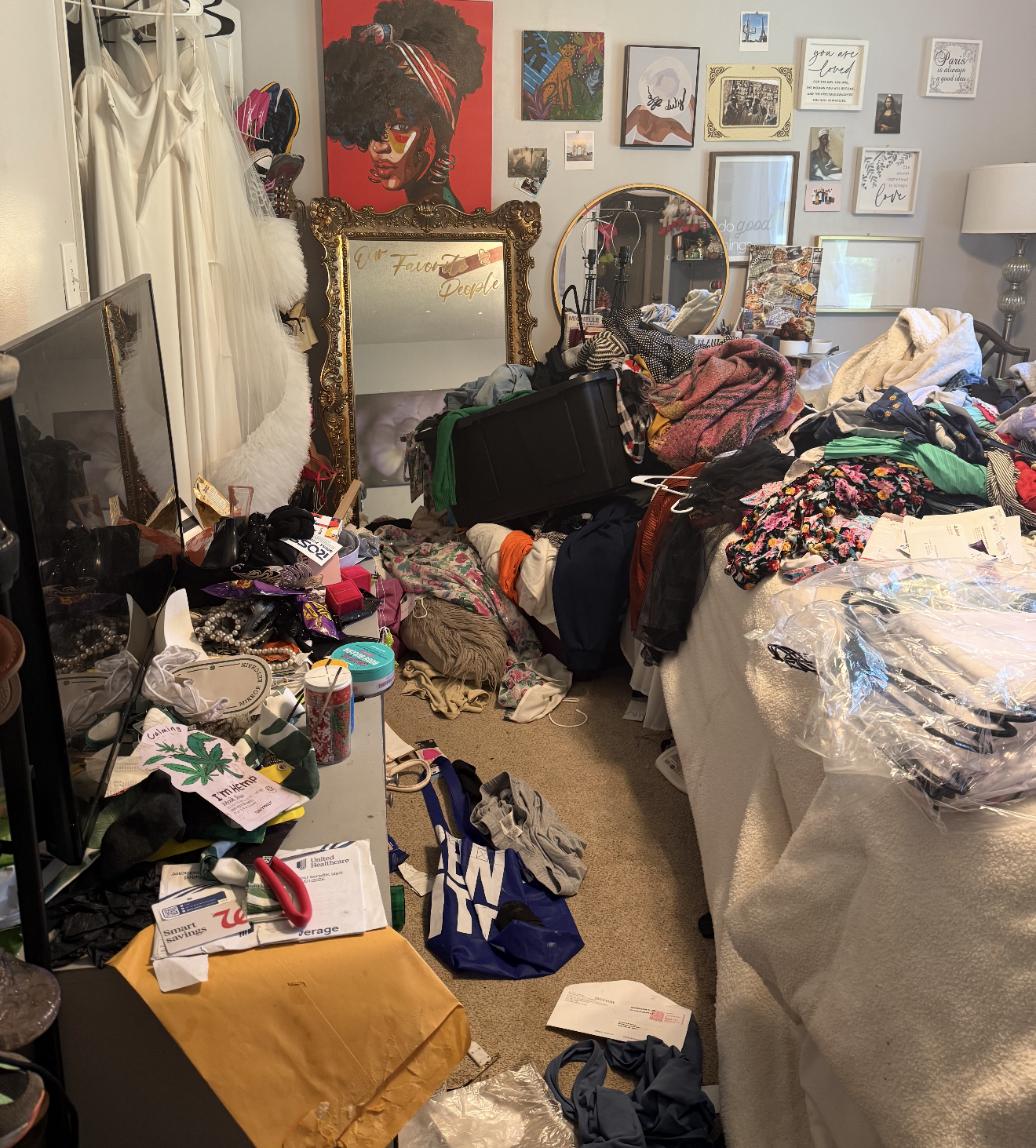 A cluttered bedroom with clothes, accessories, and miscellaneous items scattered across a bed, table, and floor. The room has wall art, including a large portrait of a woman with curly hair and sunglasses, and a variety of framed pictures and quotes. A large mirror with an ornate gold frame leans against the wall, and plastic bags containing clothing are on the bed.